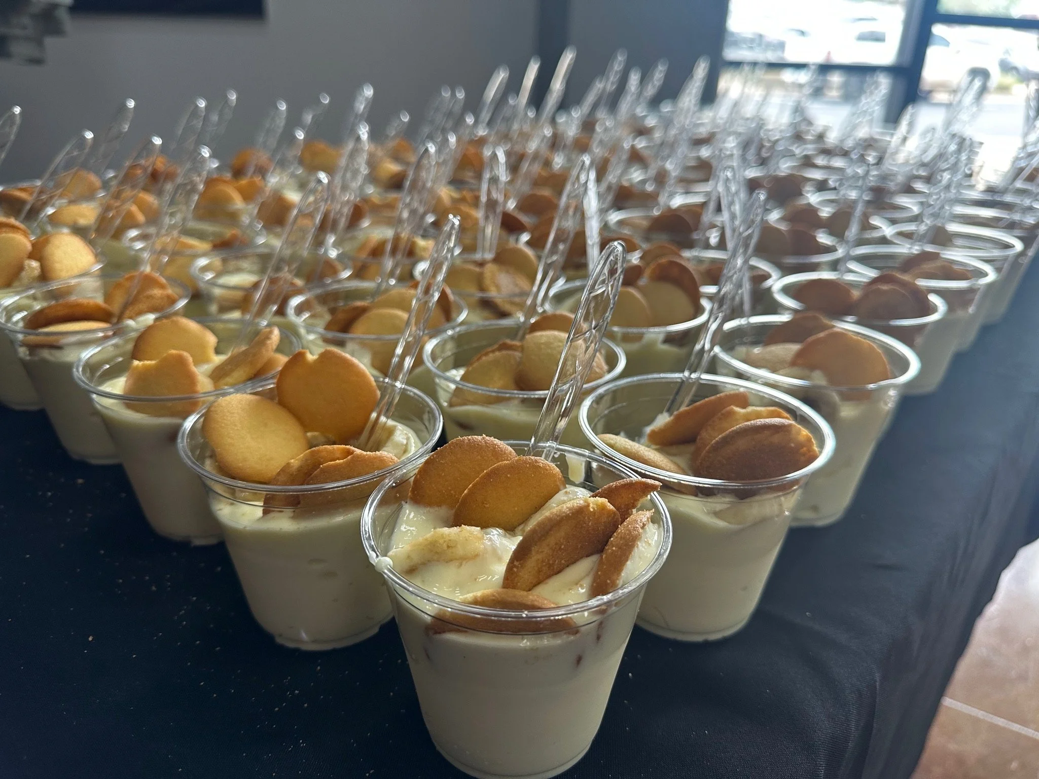 Multiple clear plastic cups filled with a layered dessert topped with cookies or wafers, lined up on a dark surface near a window.