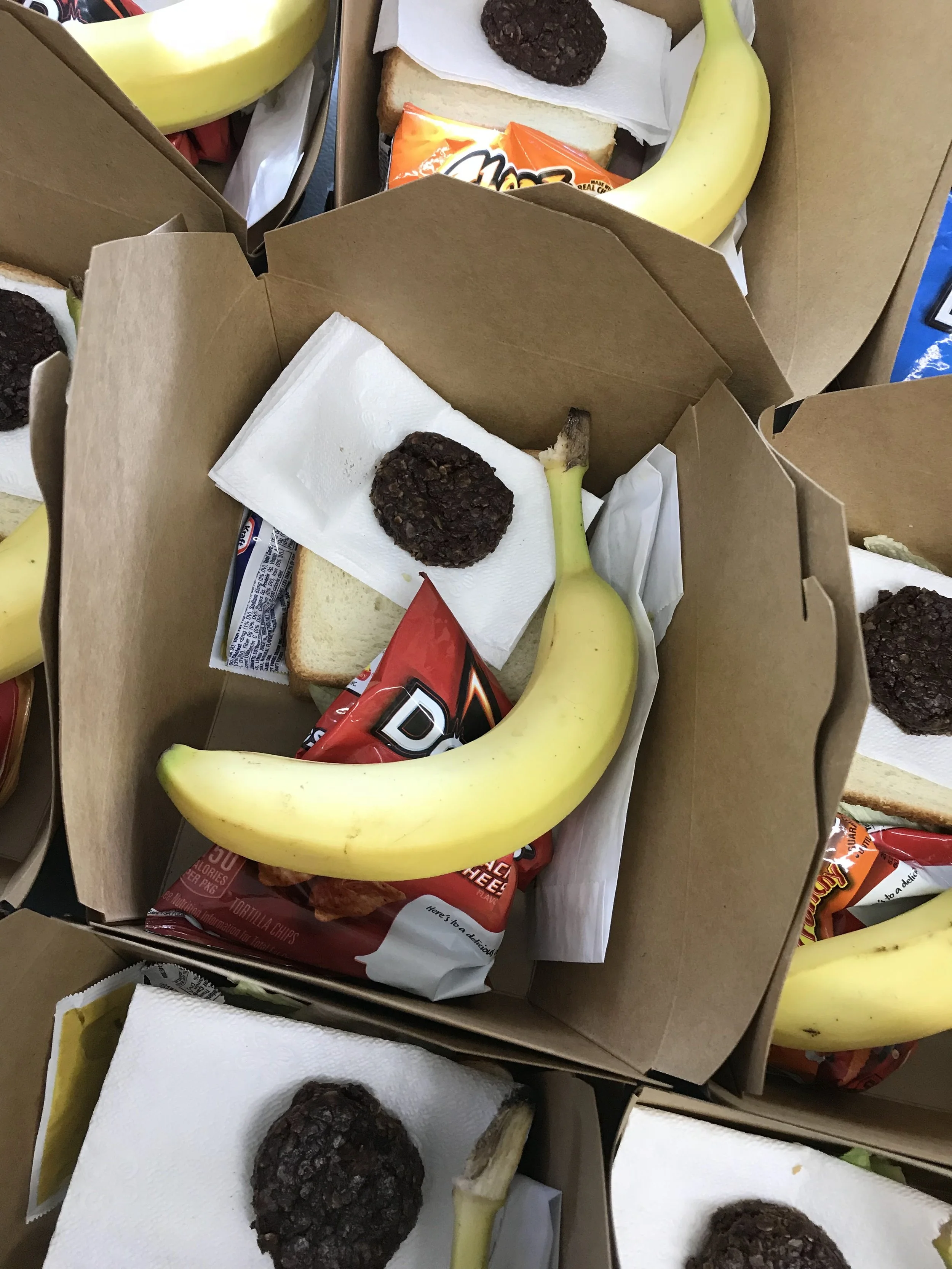 A cardboard box containing a banana, a piece of bread, chocolate cookies, and a pack of cheese crackers, lined with paper towels.