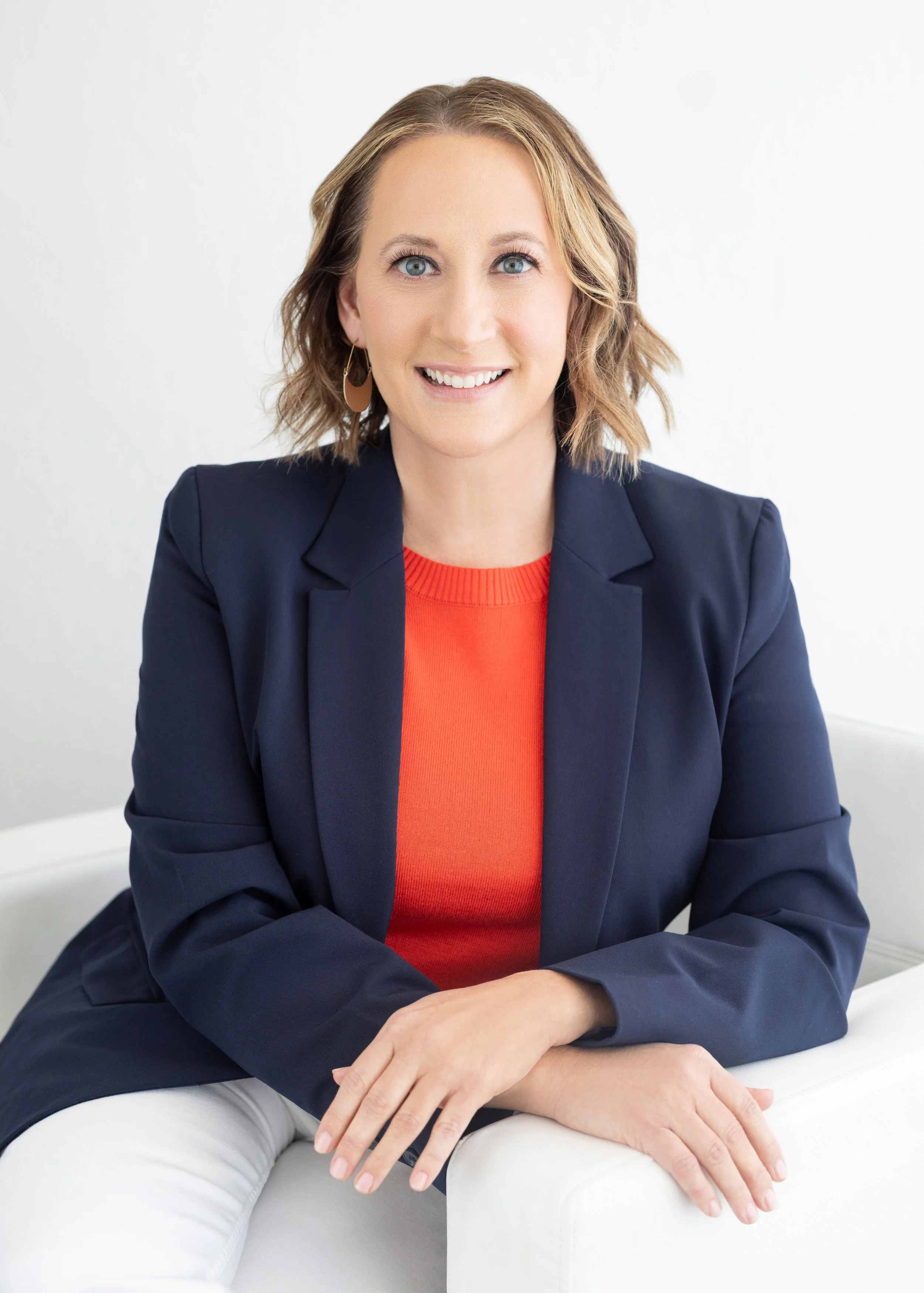 A woman with shoulder-length blonde hair, blue eyes, and light skin, smiling while sitting on a white couch. She is dressed in a navy blazer over an orange top and white pants, with gold earrings. Leadership, Team and Talent Development Coach