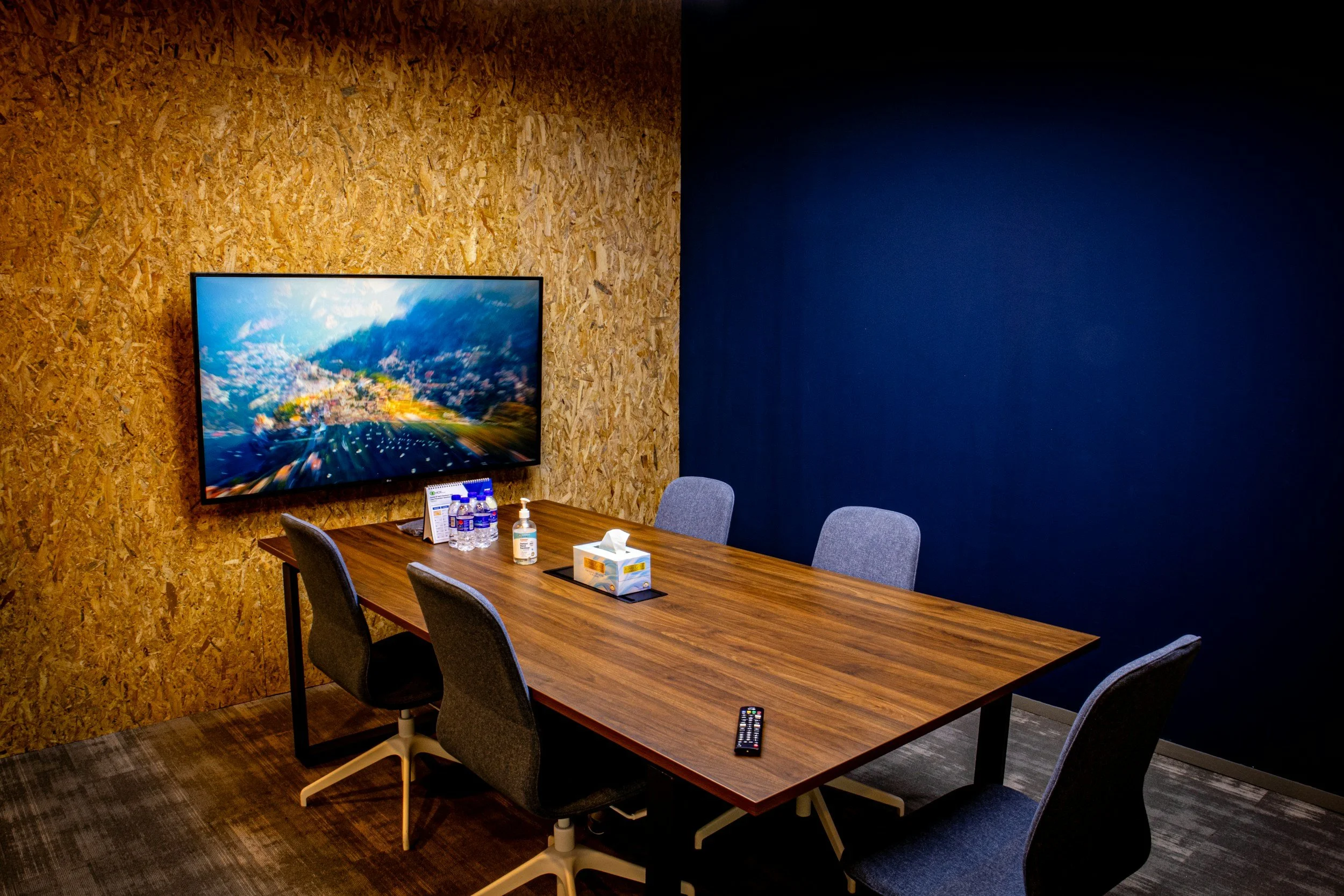 A conference room with a wooden table, six gray chairs, a wall-mounted flat screen TV displaying a scenic landscape, a remote control on the table, bottles of water, hand sanitizer, tissues, and a few small items.
