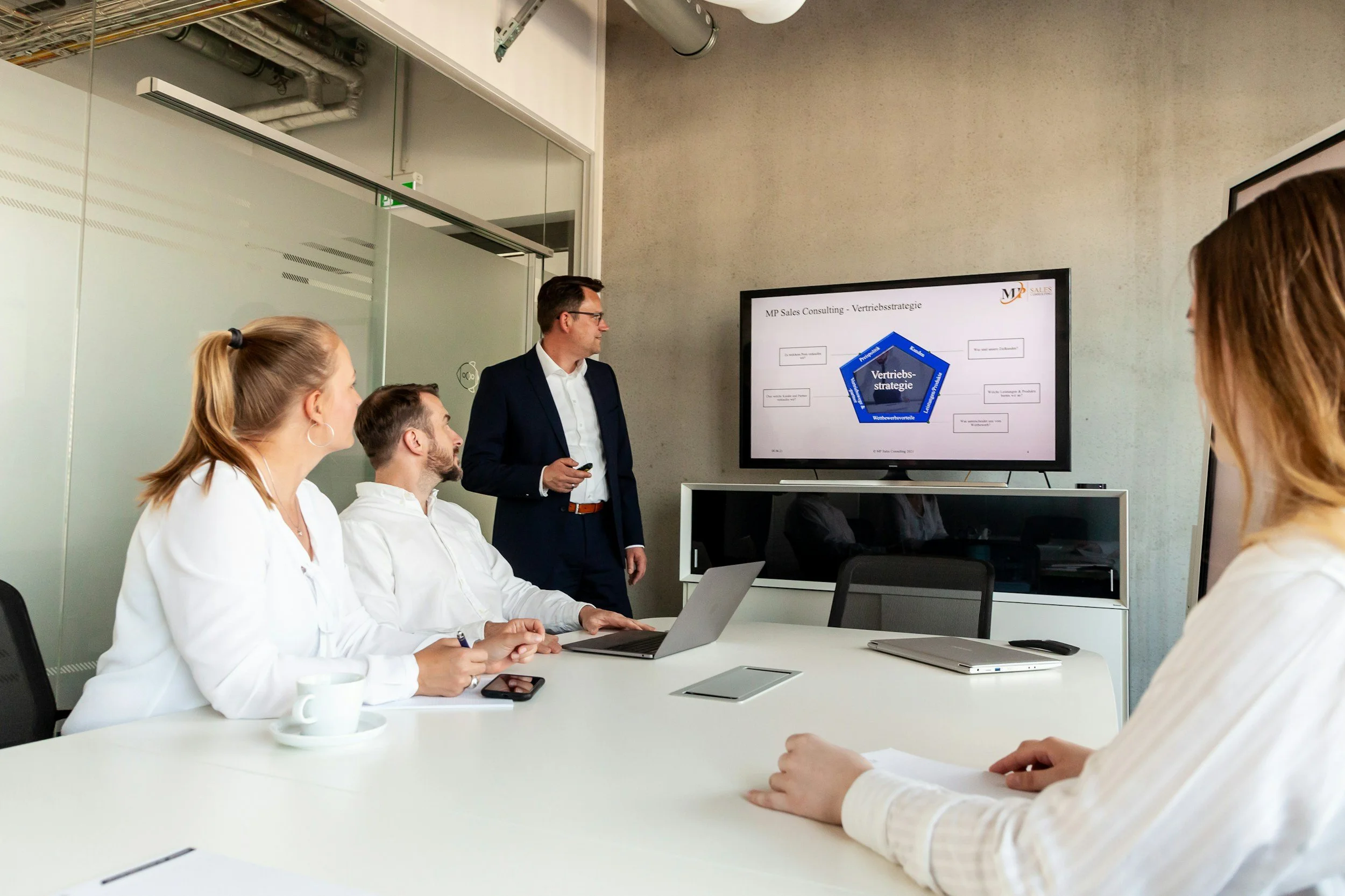 A man in a suit giving a presentation to a group of four women in a modern conference room, with a large screen displaying a sales strategy diagram.