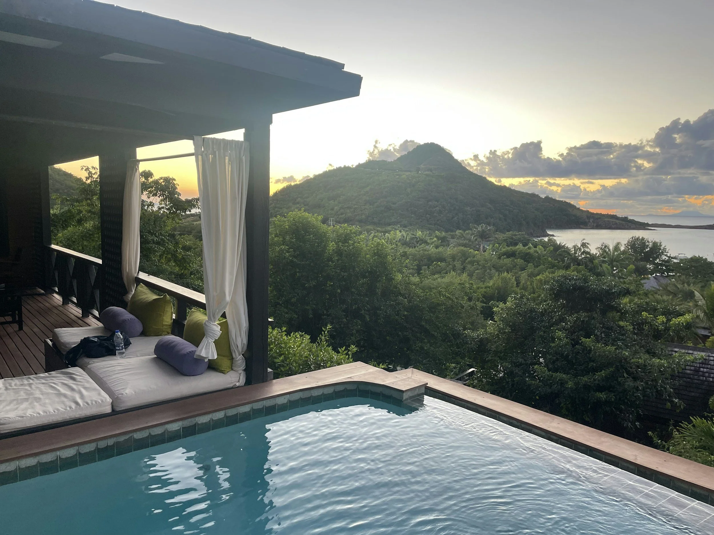View of a swimming pool with a mountain and lush green landscape in the background during sunset, with curtains hanging on a deck and outdoor lounge furniture.