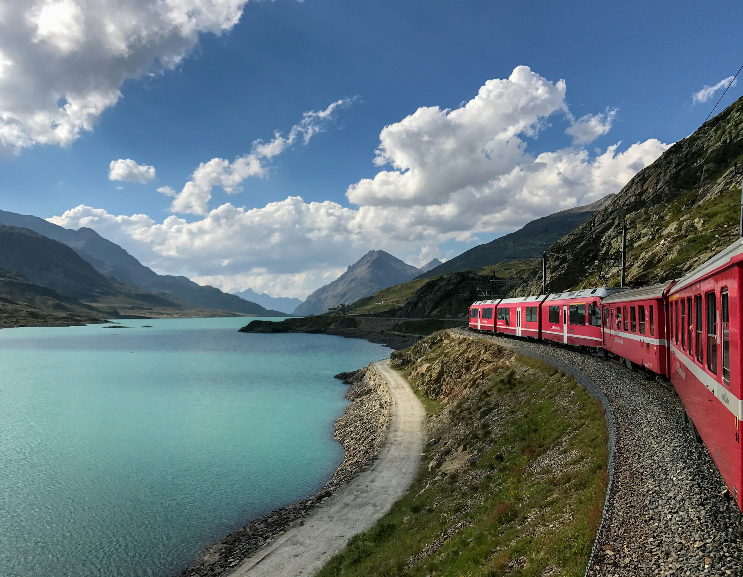 A red train traveling along a track beside a turquoise lake with mountains under a partly cloudy sky.