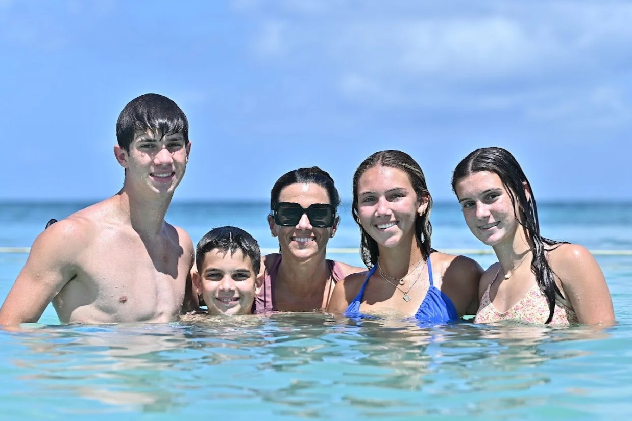 A group of five people, including two adult women and three children, smiling and standing in the shallow ocean water with a bright blue sky and ocean in the background.