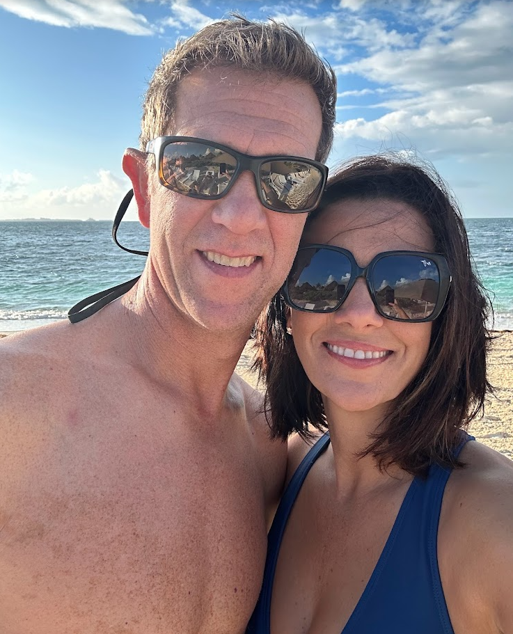 A man and woman taking a selfie on the beach during the day, both wearing sunglasses and smiling, with the ocean and partly cloudy sky in the background.