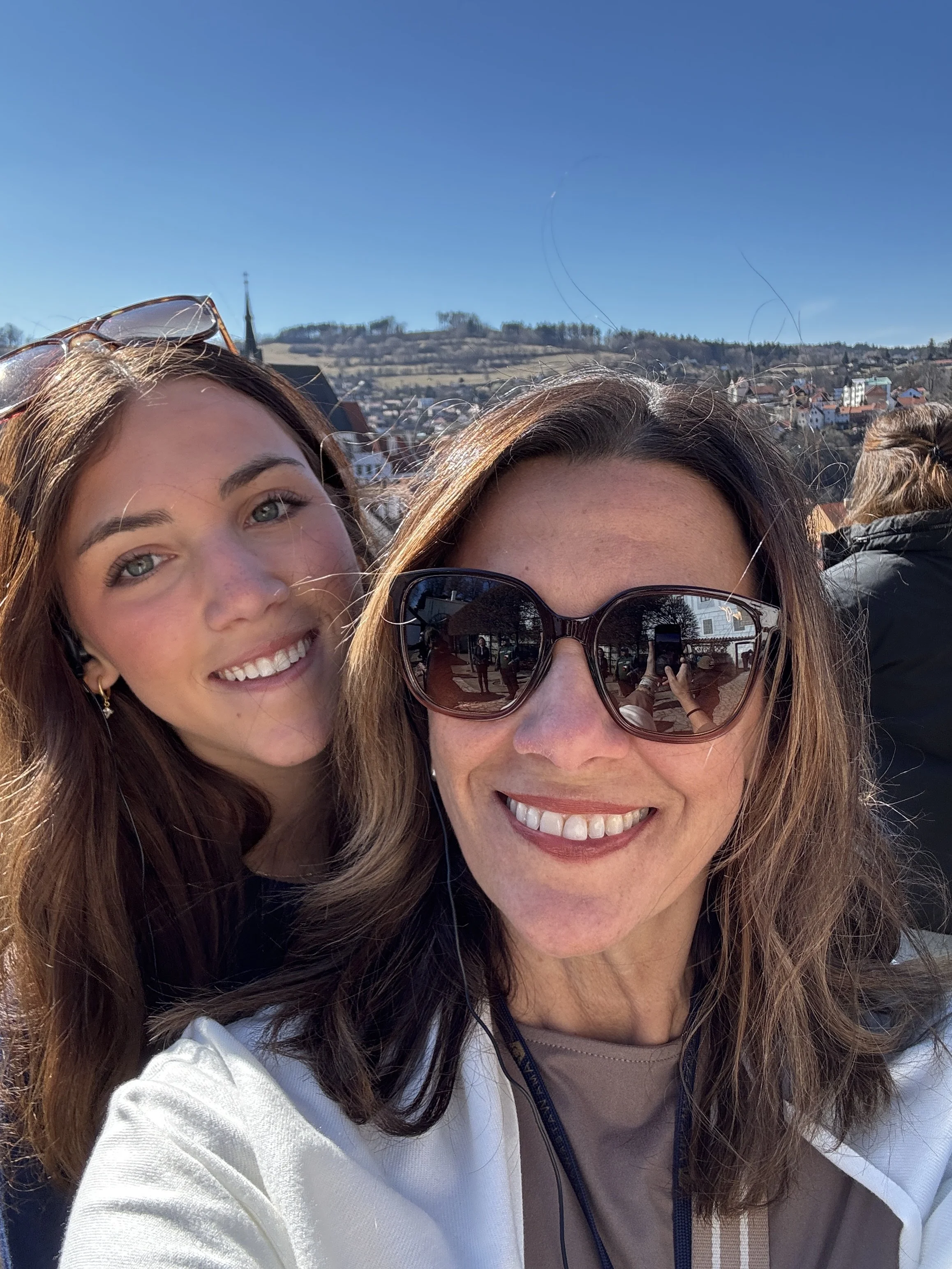 Two women smiling outdoors on a sunny day, with a scenic background of hills and houses.