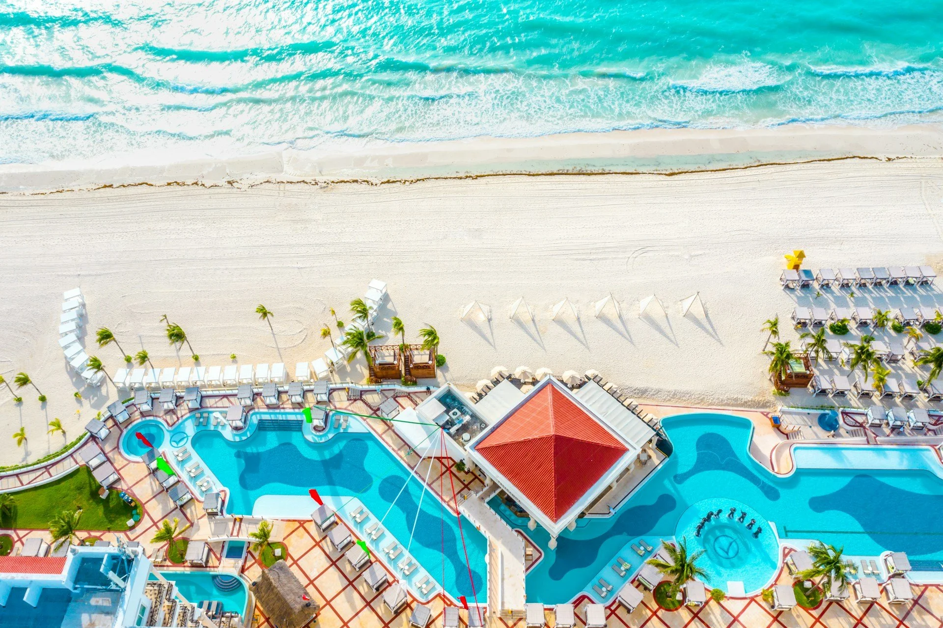 Aerial view of a beach resort showing a sandy beach, ocean waves, pool area with lounge chairs, palm trees, and a building with a red roof.