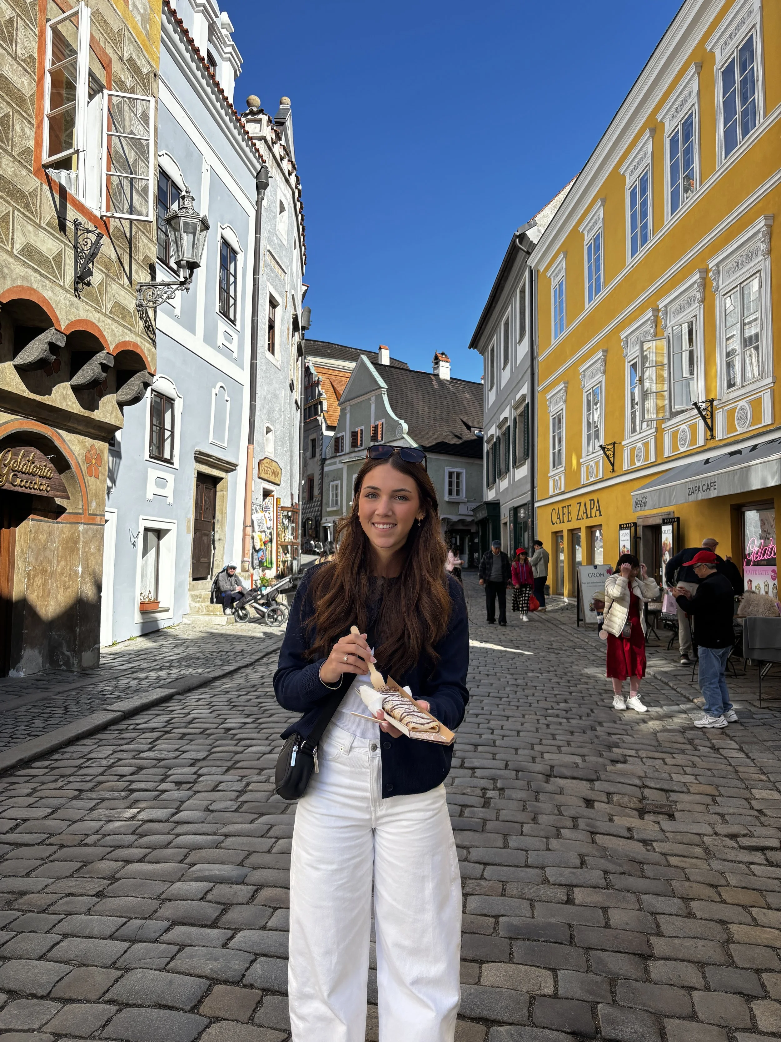 A young woman with long brown hair, sunglasses on her head, wearing a black jacket and white pants, is standing on a cobblestone street holding a tray with a dessert. Behind her, there are several colorful buildings with European architecture, including a yellow building labeled "CAFE ZAPA," and various people walking and chatting.
