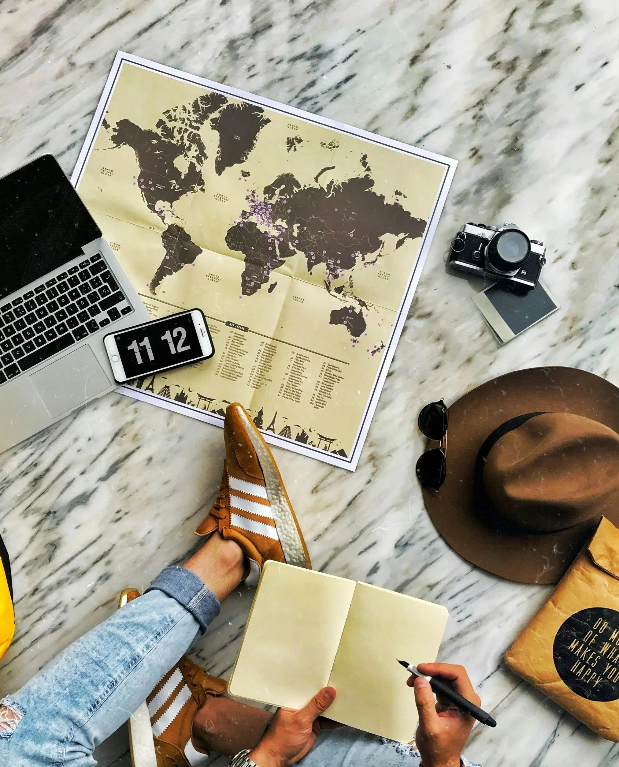 A busy person or mom wearing ripped jeans and brown shoes is sitting on a marble floor, writing in a yellow notebook with a black pen. There is a map of the world, a camera, sunglasses, a smartphone, and a brown bag nearby. planning a vacation.