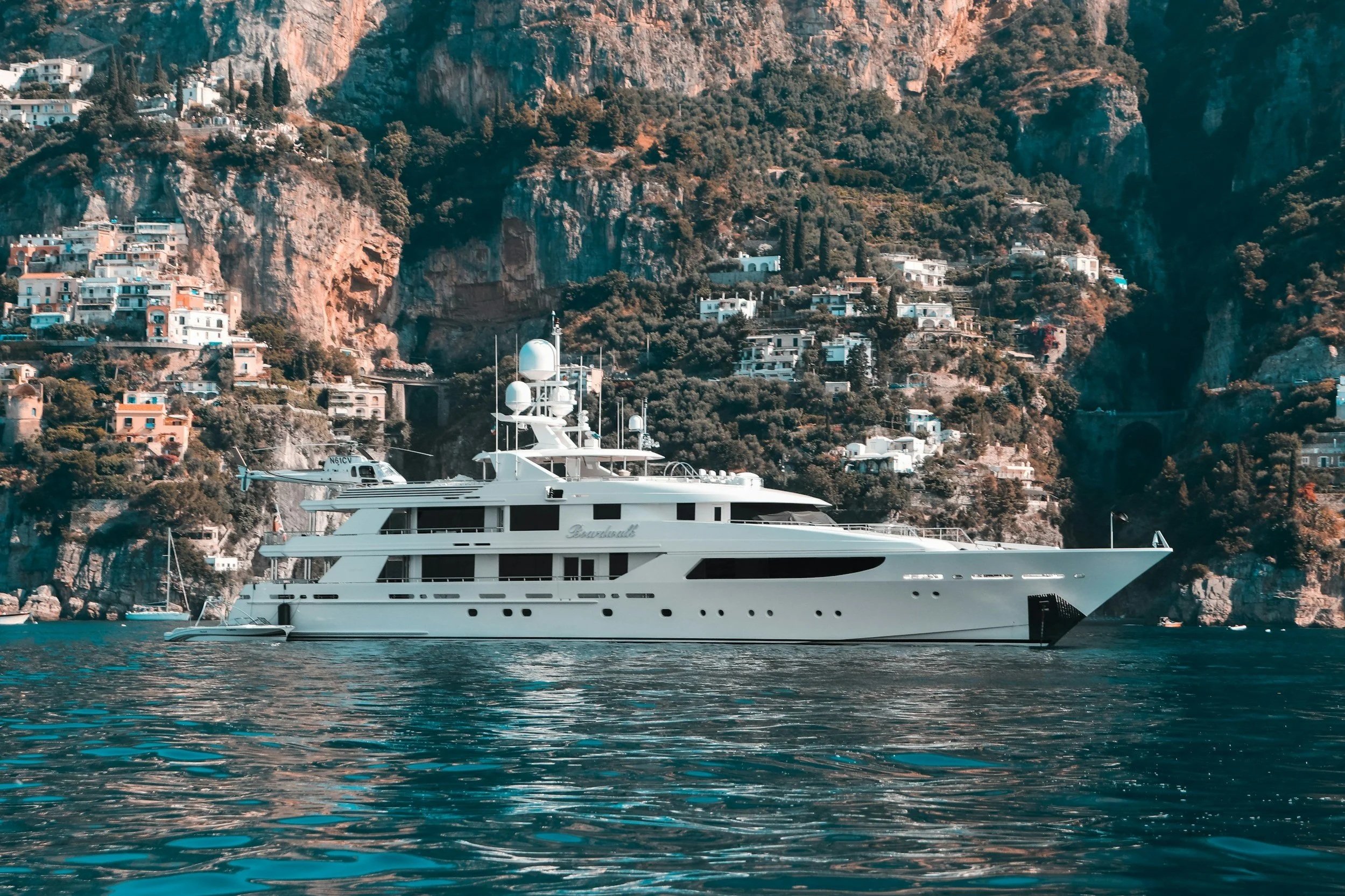 Large white yacht with multiple decks floating in water near a steep hillside with numerous houses and greenery.