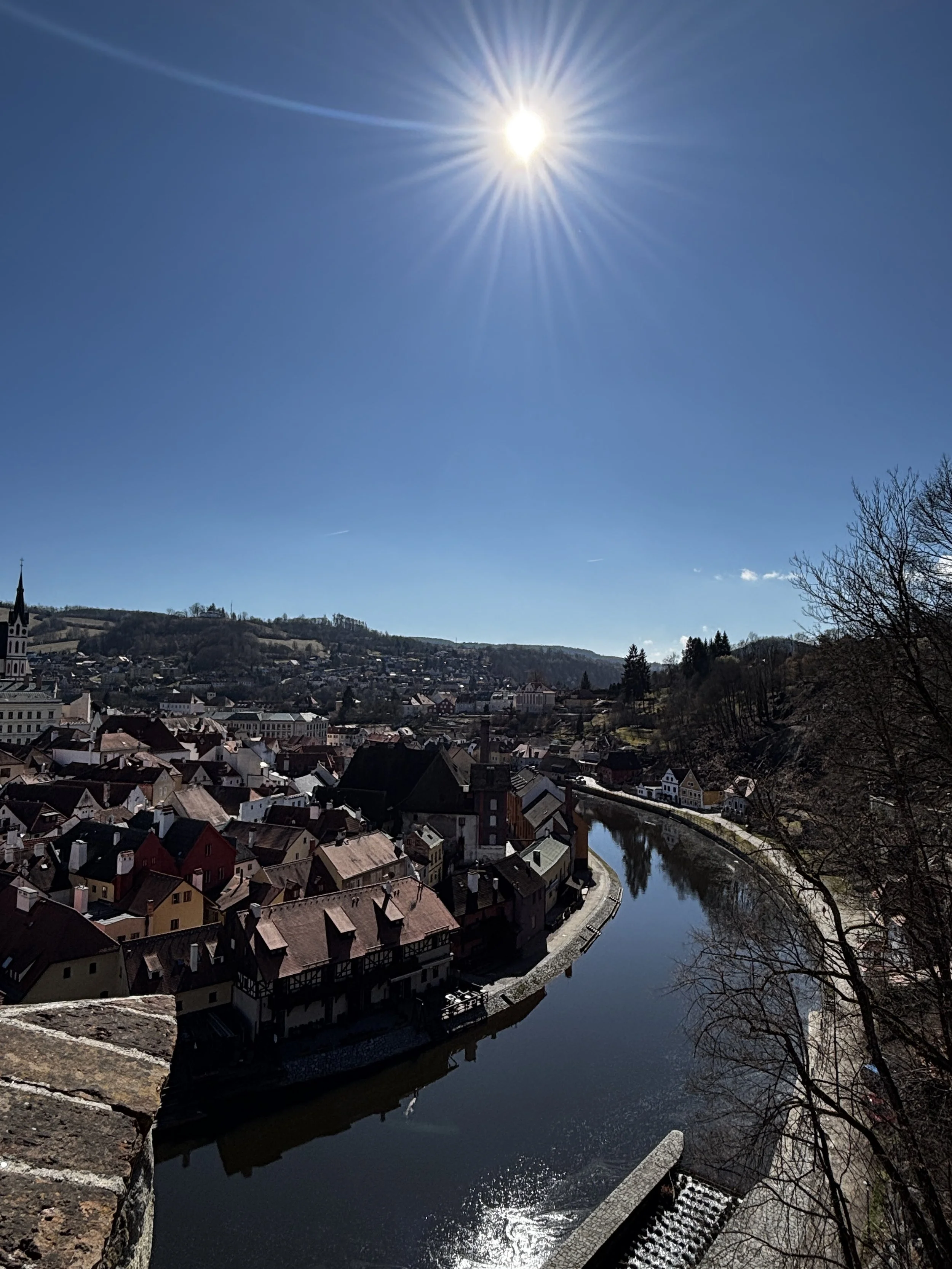 A scenic view of a town by a river, Cesky Krumlov, a stop on a European River Cruise with a hill in the background, under a bright sun in a clear blue sky.