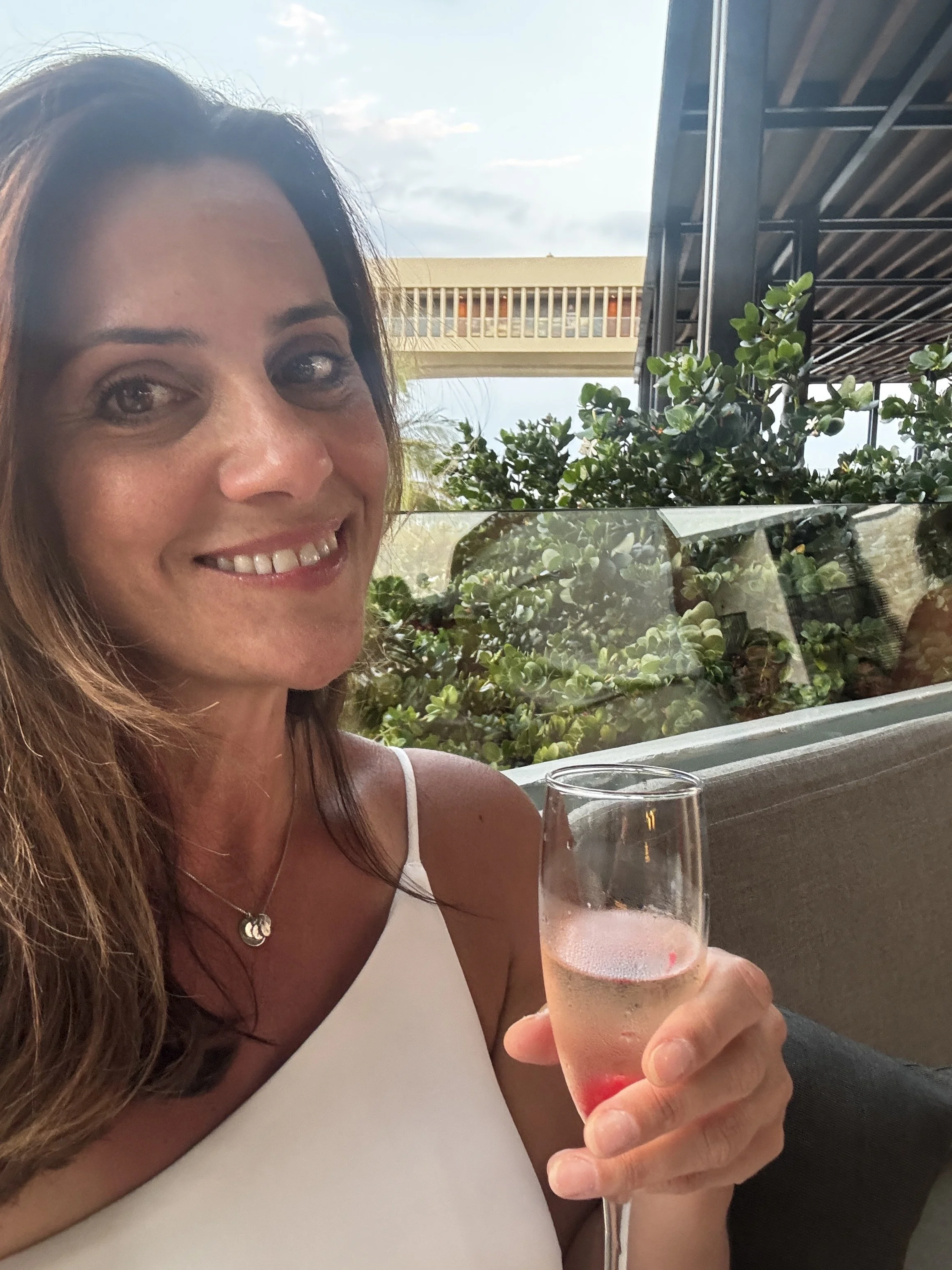 Nikki Trefzger, Travel by Nikki Vacations smiling and holding a glass of rosé wine at an all inclusive resort in the Caribbean, with greenery and a building with a balcony in the background.