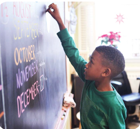A young boy writing on a blackboard in a classroom with a flower decoration and sunlight coming through the window.