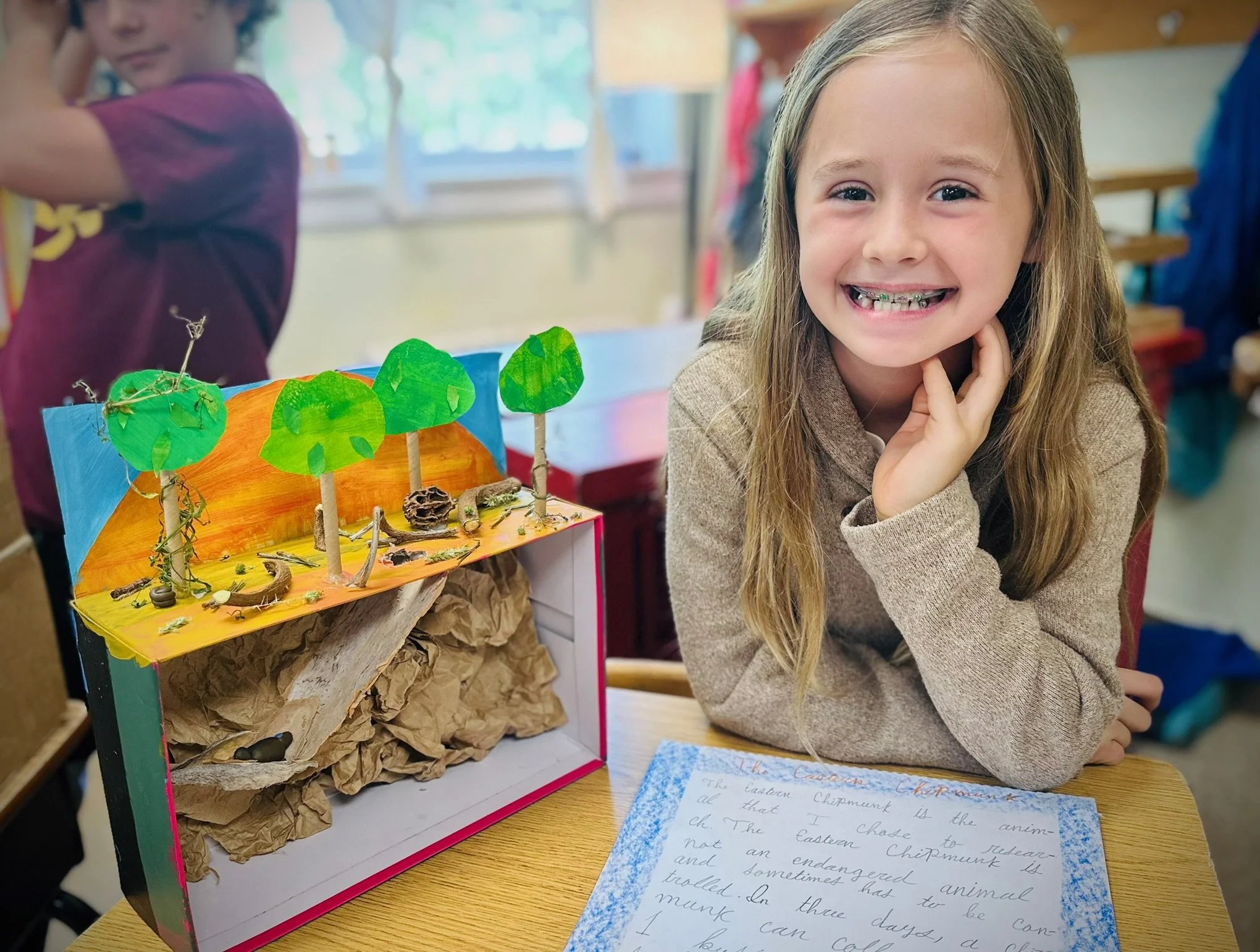 A young girl smiling with braces in a classroom, sitting at a desk next to a colorful diorama of trees and a landscape, with a handwritten notebook in front of her.