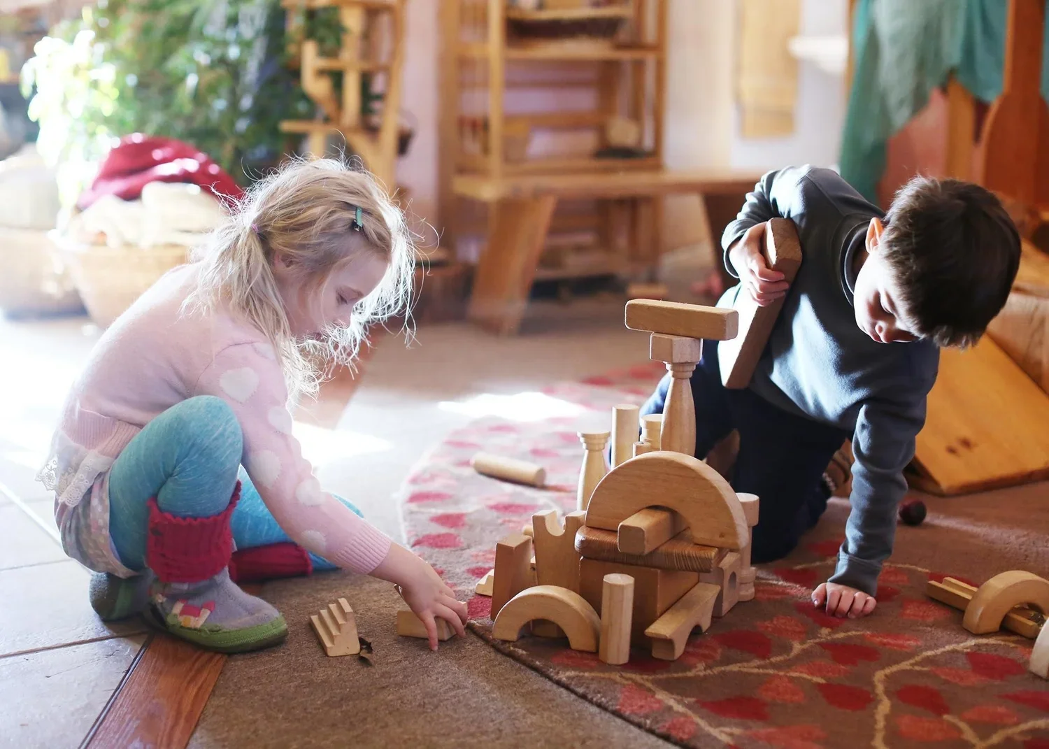 Two children, a girl and a boy, are playing with wooden building blocks on the floor indoors. The girl is sitting cross-legged and wearing colorful socks and pajamas, while the boy is kneeling and dressed in a dark shirt. They are constructing a structure with the blocks.