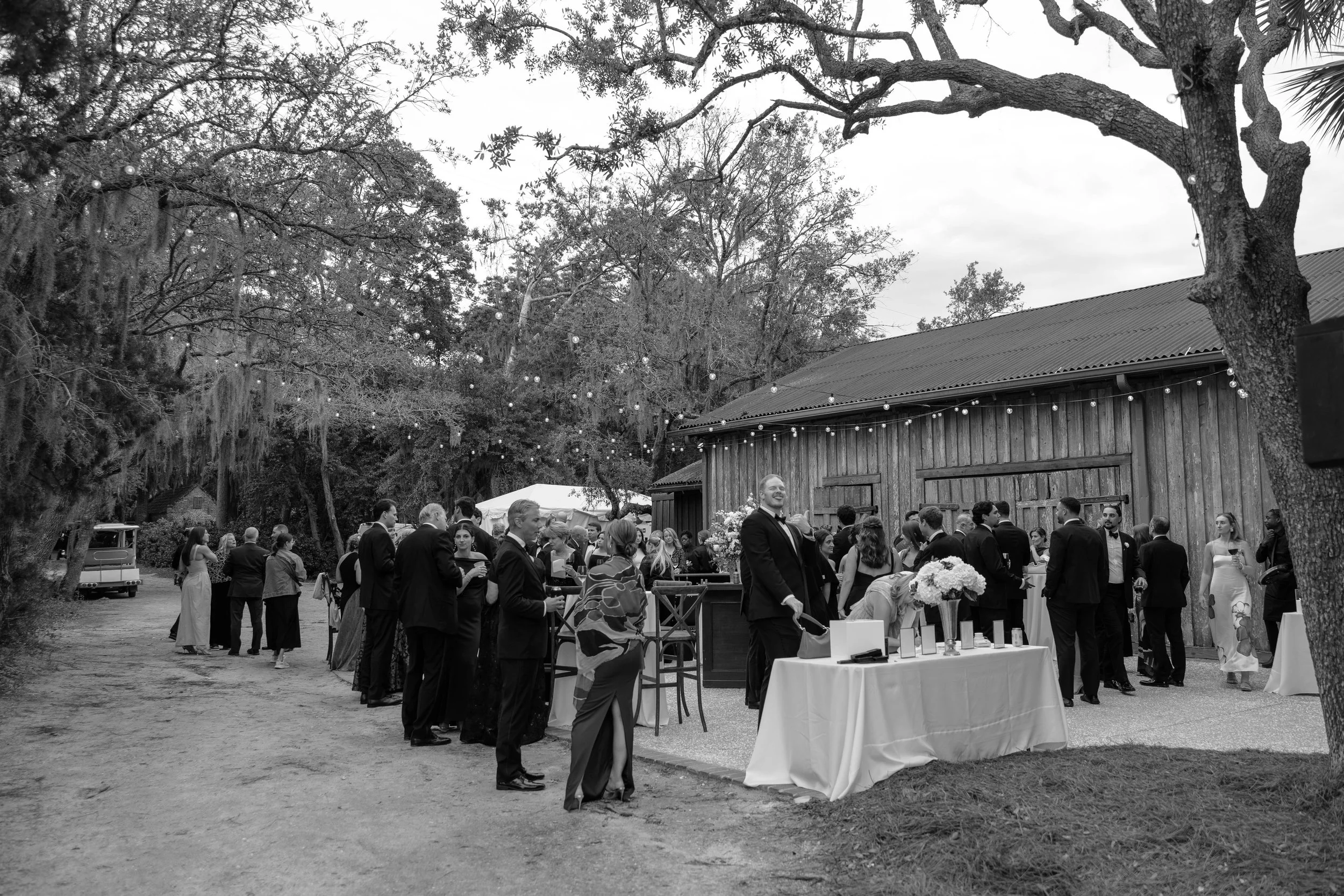 People gathered outdoors for a celebration or event, standing behind a table with props, surrounded by trees and a rustic building, with string lights hanging above.