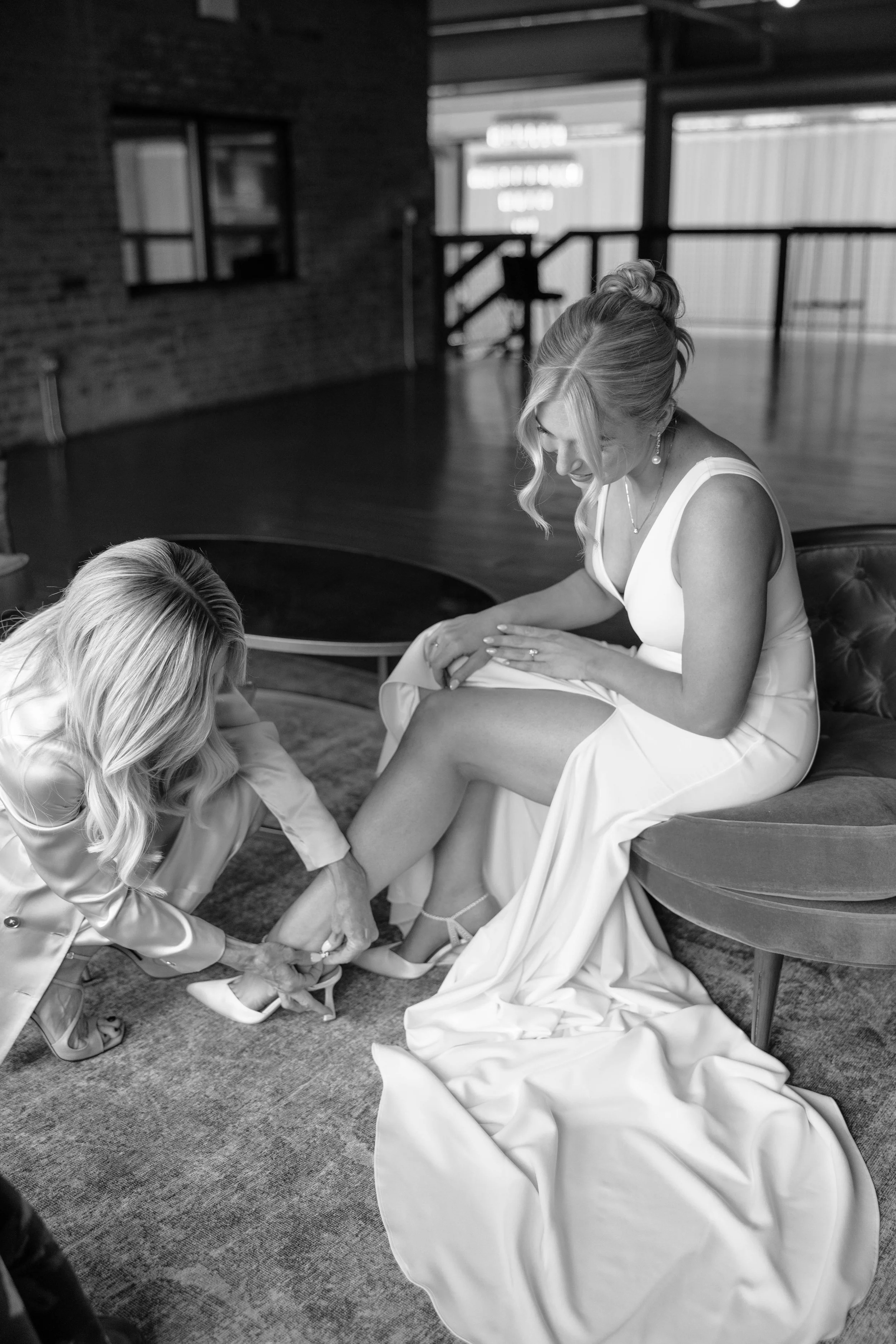 A woman in a wedding dress sits in a chair while another woman helps her put on a shoe. The scene is indoors with natural light coming through windows.