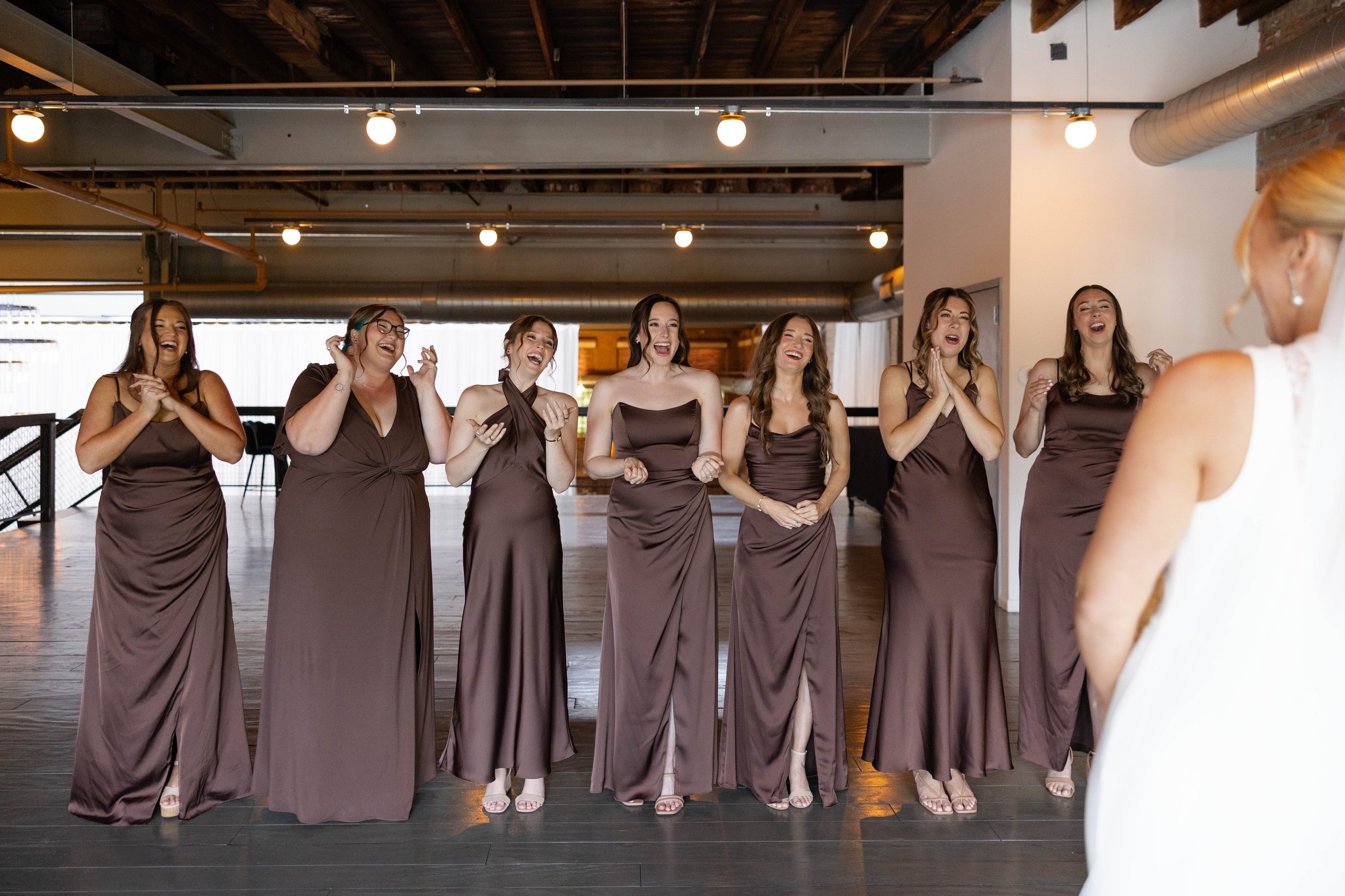 A group of seven women in matching brown dresses laughing and smiling at a woman in a white dress, in a modern indoor setting with wooden floors and exposed ceilings.