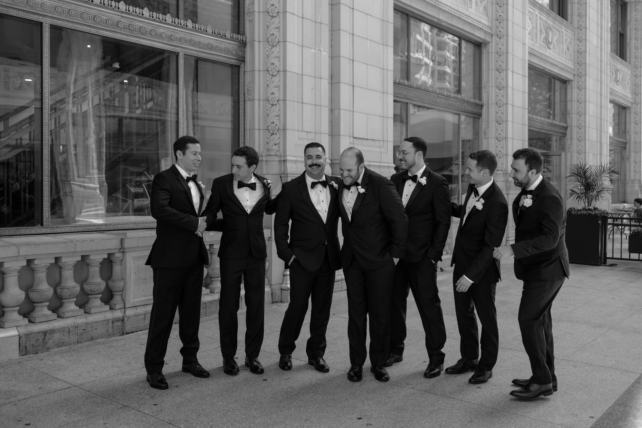 A group of seven men dressed in tuxedos, standing outside a building with ornate architectural details, smiling and socializing.