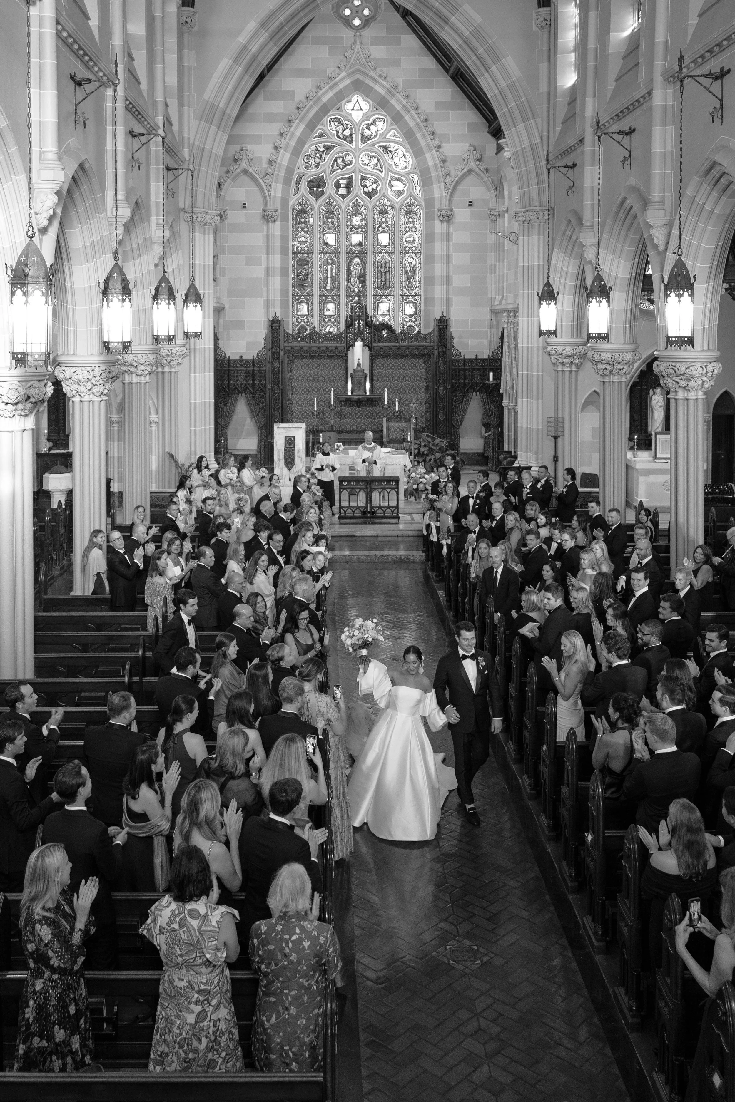 A wedding ceremony inside a church with many attendees, a bride and groom walking down the aisle, and a priest officiating the ceremony.