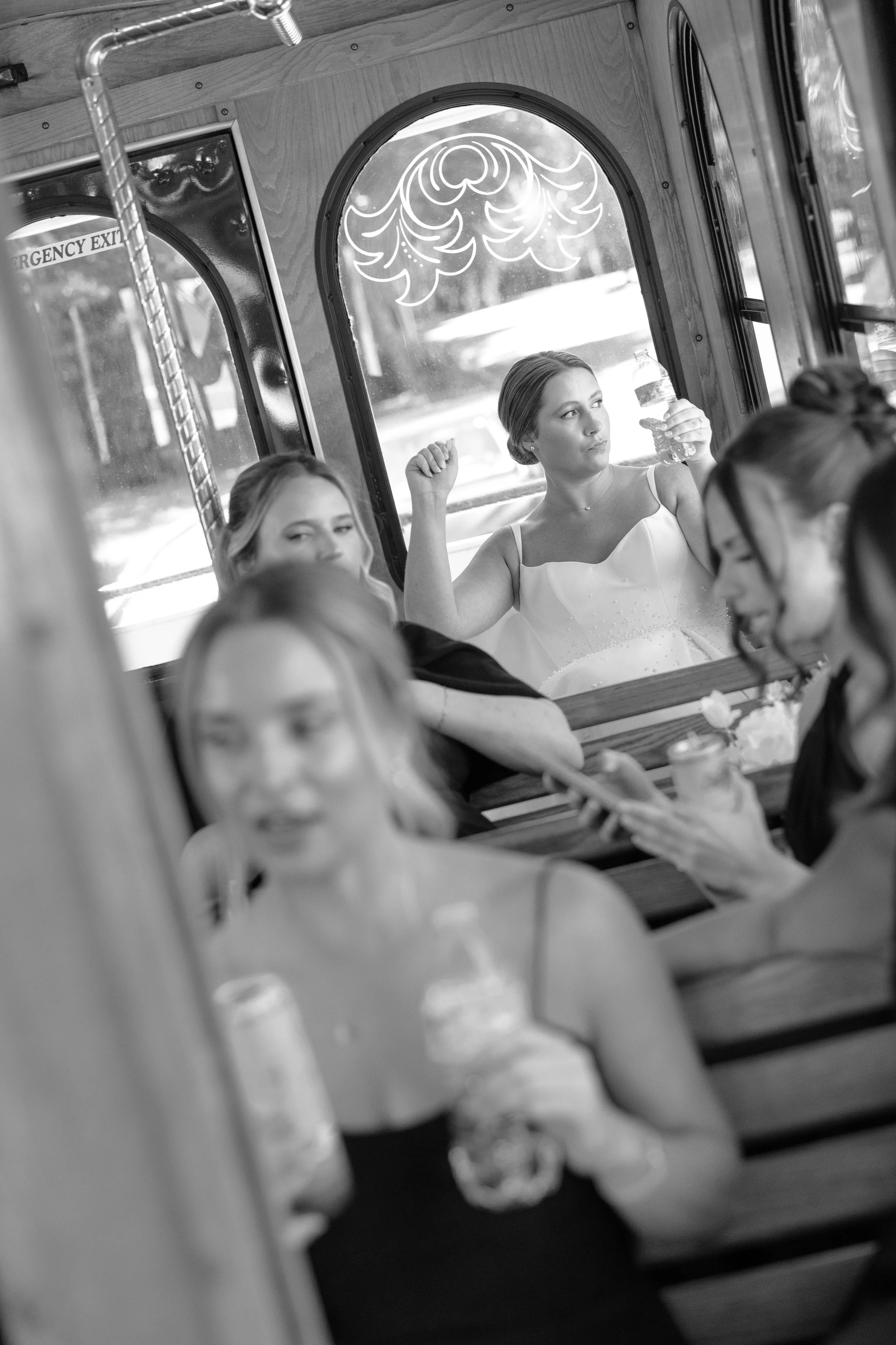 Several women sitting in a bus, one holding a water bottle, others engaged in conversation or looking around. The bus interior has wooden paneling and decorative window trim.