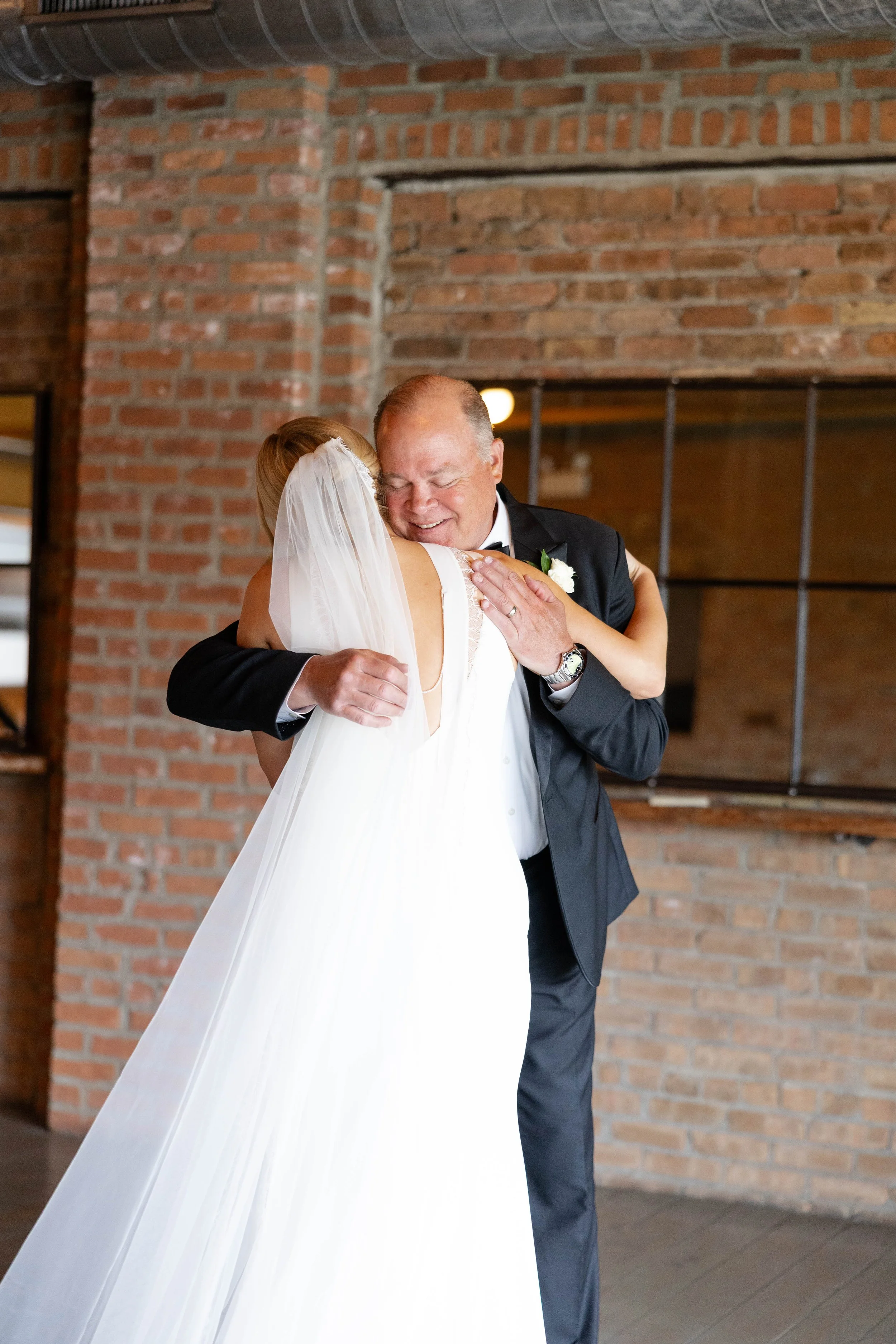 A bride and a groom embracing each other on their wedding day, with the bride in a white dress and veil, and the groom in a black tuxedo, in front of a brick wall.