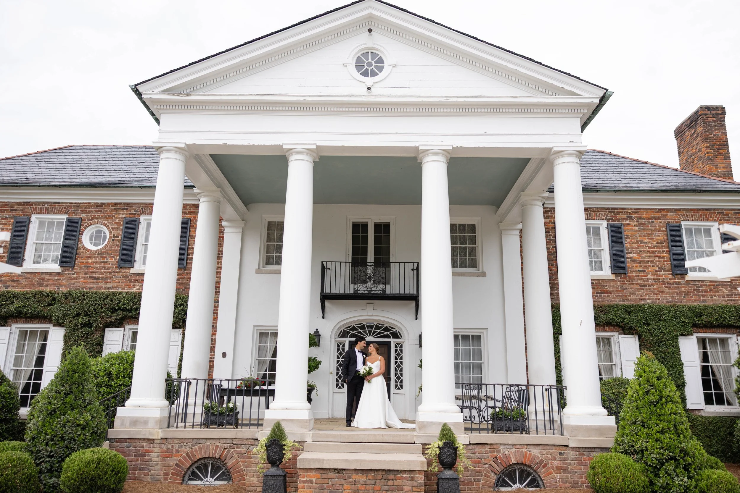 A bride and groom standing on the steps of a large white mansion with tall columns, brick walls, and multiple windows, holding hands and looking at each other.