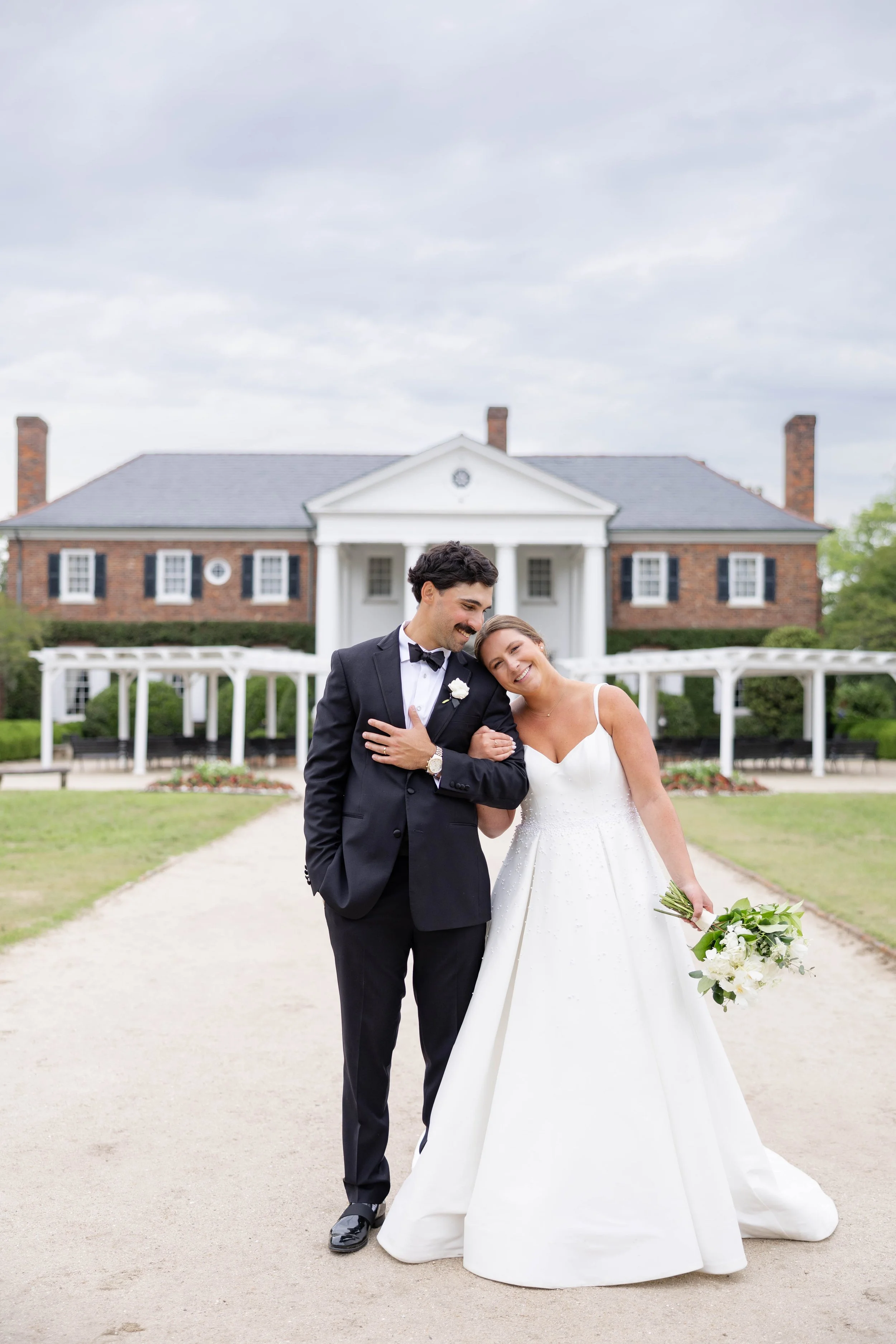 A bride and groom standing outdoors in front of a large, historic brick house with white columns and a garden. The bride is holding a bouquet of white flowers and leaning her head on the groom's shoulder. The groom is dressed in a black tuxedo with a