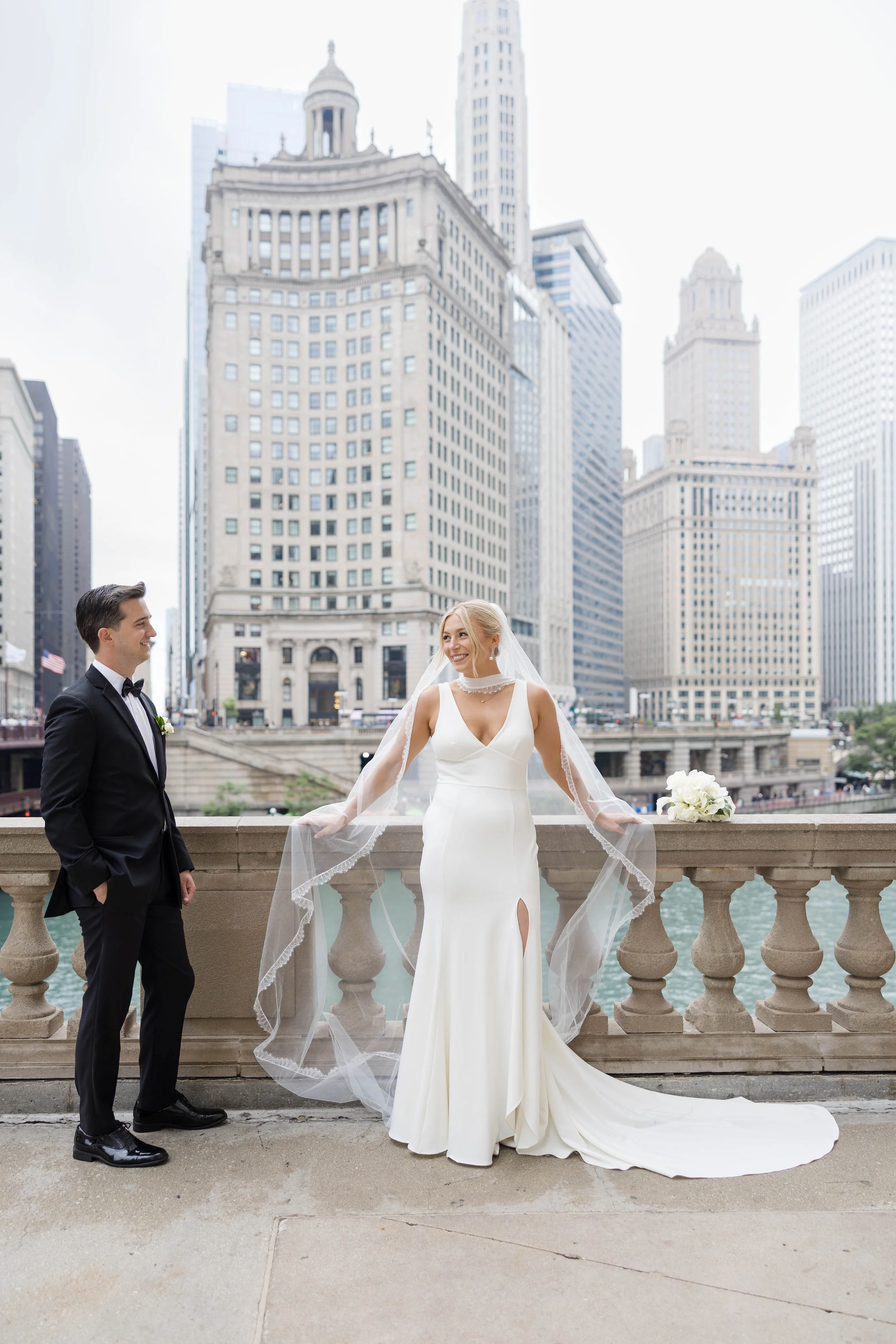 A bride in a white wedding dress and veil standing by a stone railing with a bouquet of white flowers, smiling at a groom in a tuxedo with a bow tie, on a bridge with a cityscape of tall buildings in the background.
