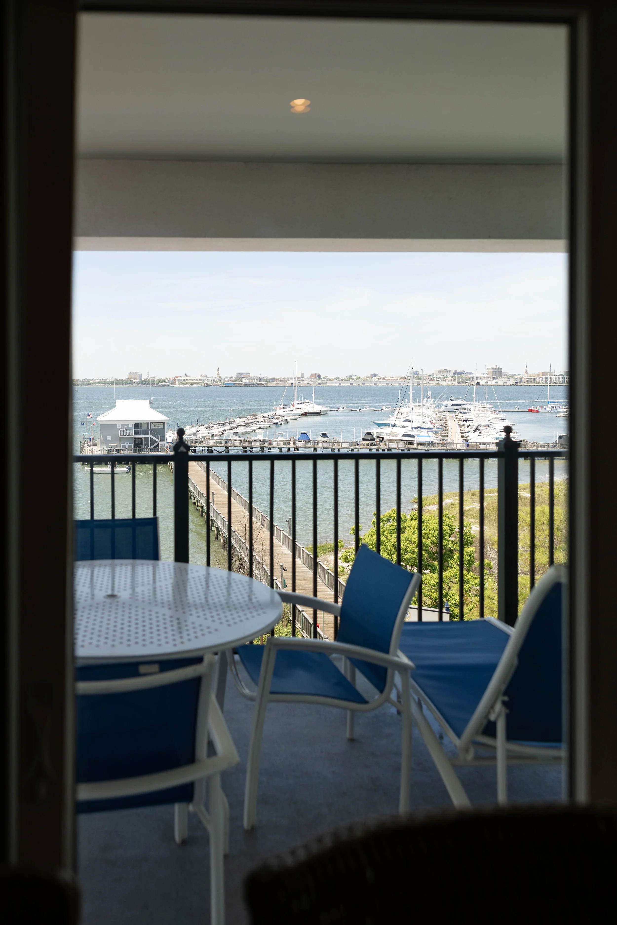 Balcony view of a marina with boats and a distant city skyline, seen from a patio with blue chairs and a white table.