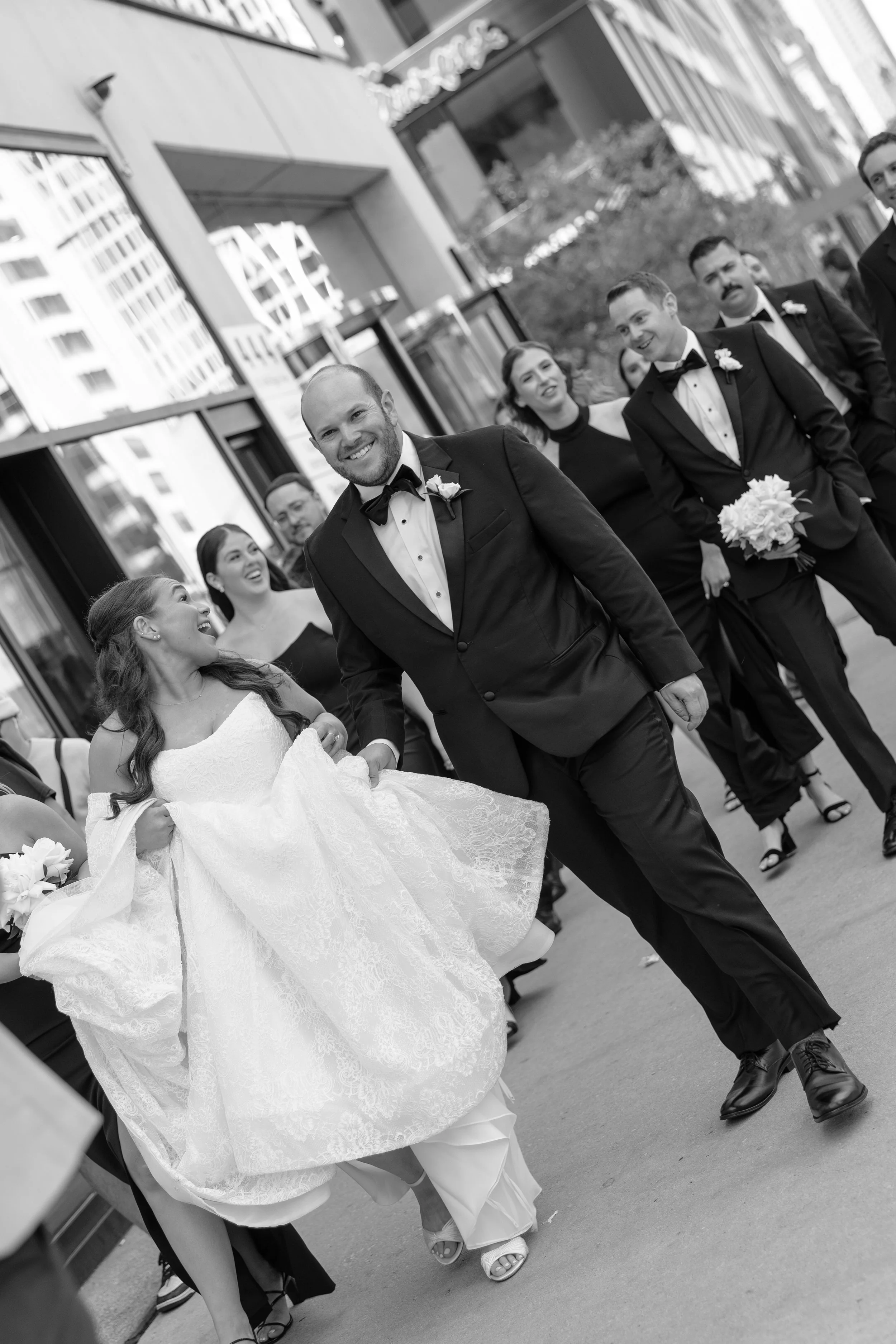 Black and white photo of a wedding procession walking outside in an urban area, with the bride in a white lace wedding dress holding a bouquet, and the groom in a tuxedo, smiling. Several bridesmaids and groomsmen accompany them, dressed formally, wa