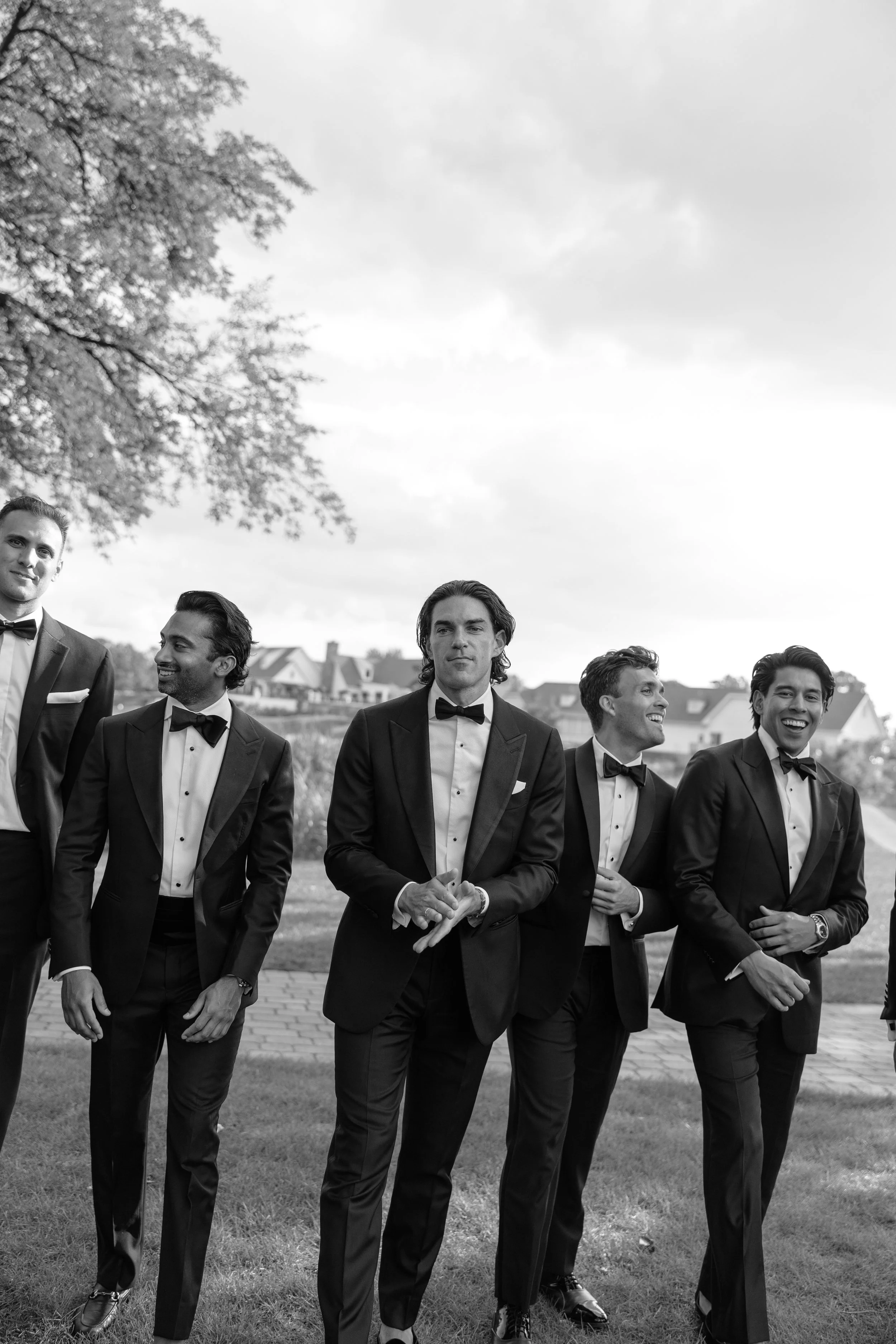 Group of men in tuxedos walking outdoors on a cloudy day, smiling and talking to each other.