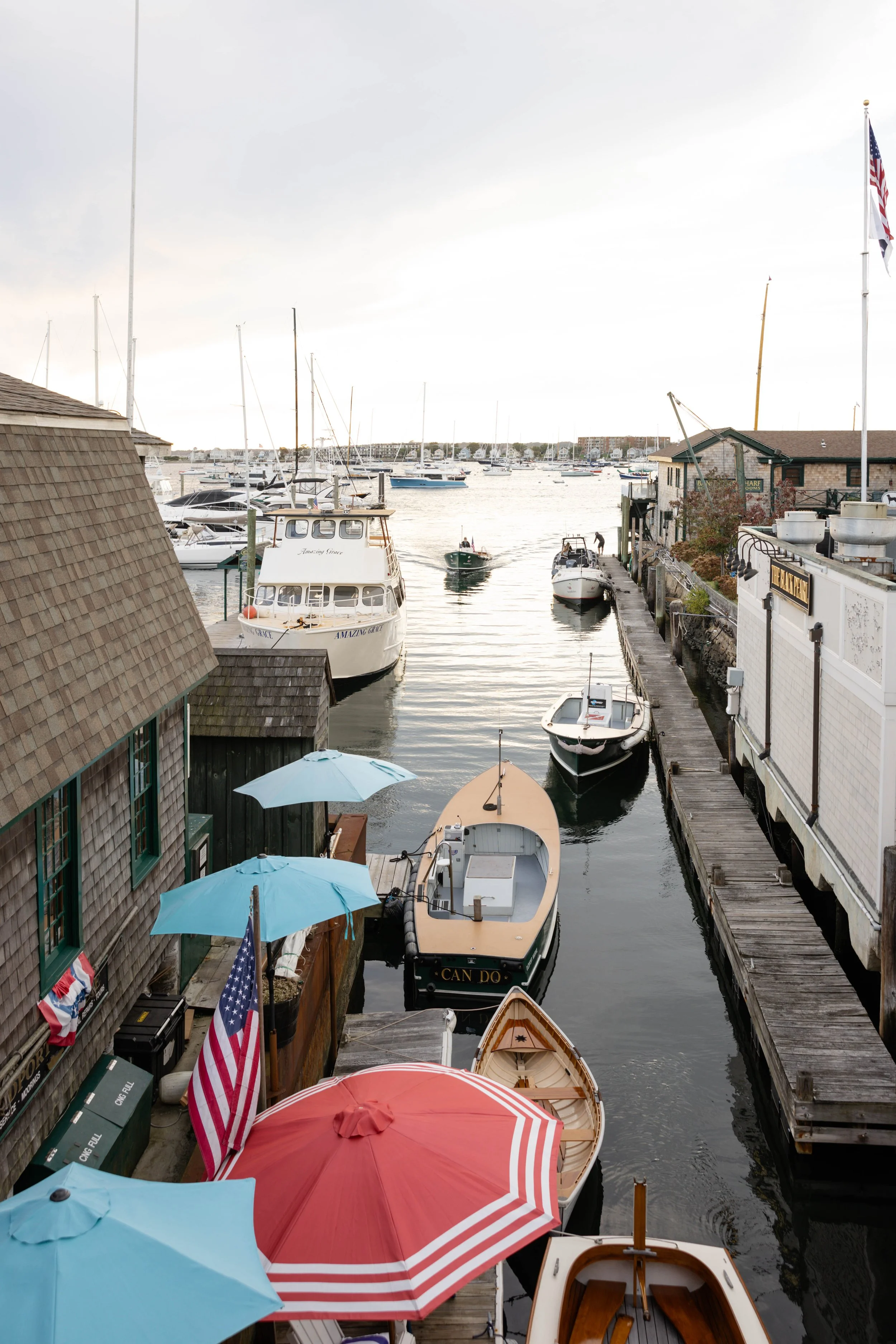 View of a marina with multiple boats docked along wooden piers, colorful umbrellas on the dock, and a cloudy sky overhead.