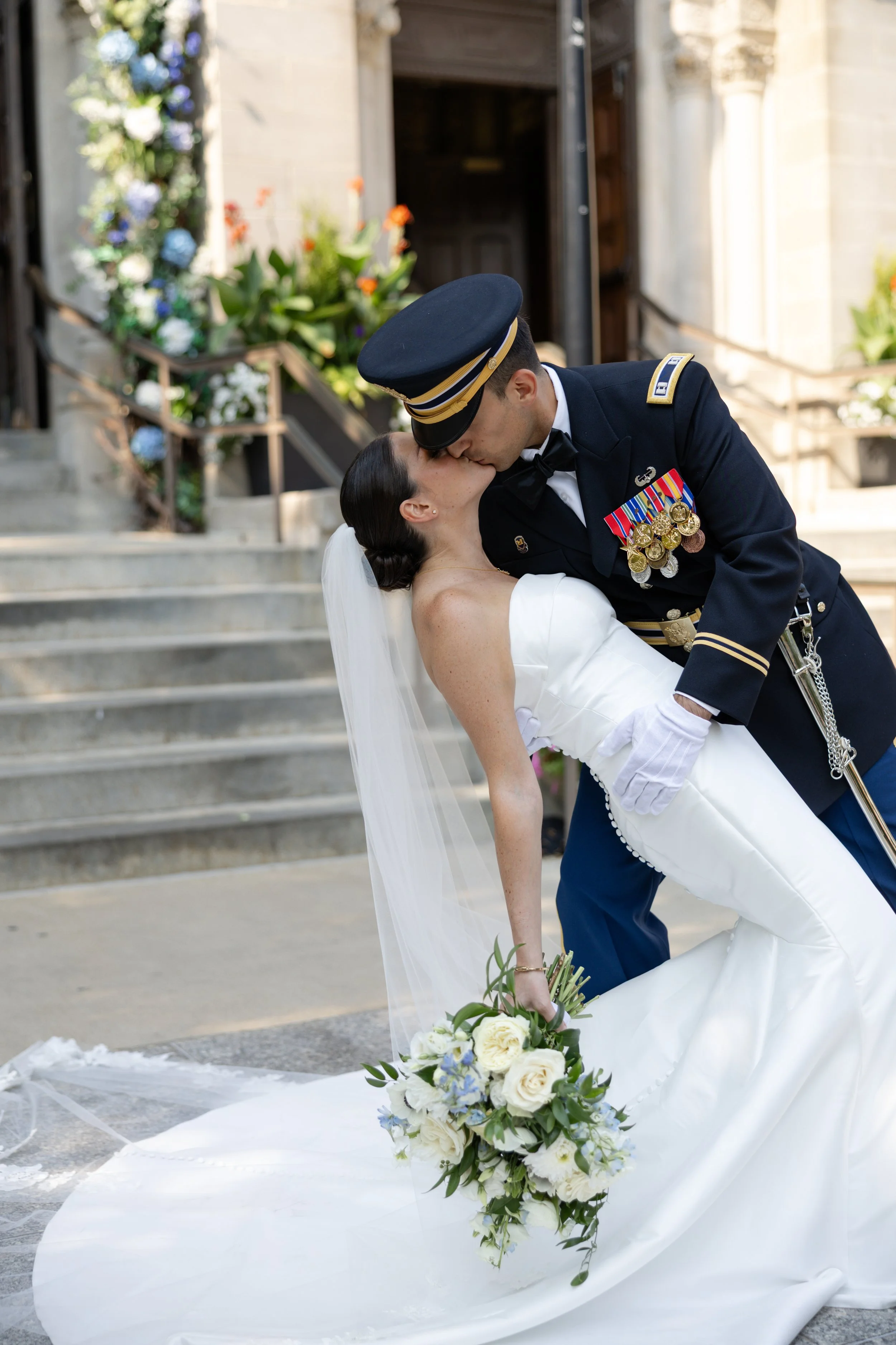 A bride and a groom in military uniform kissing outside a building with steps and flowers.