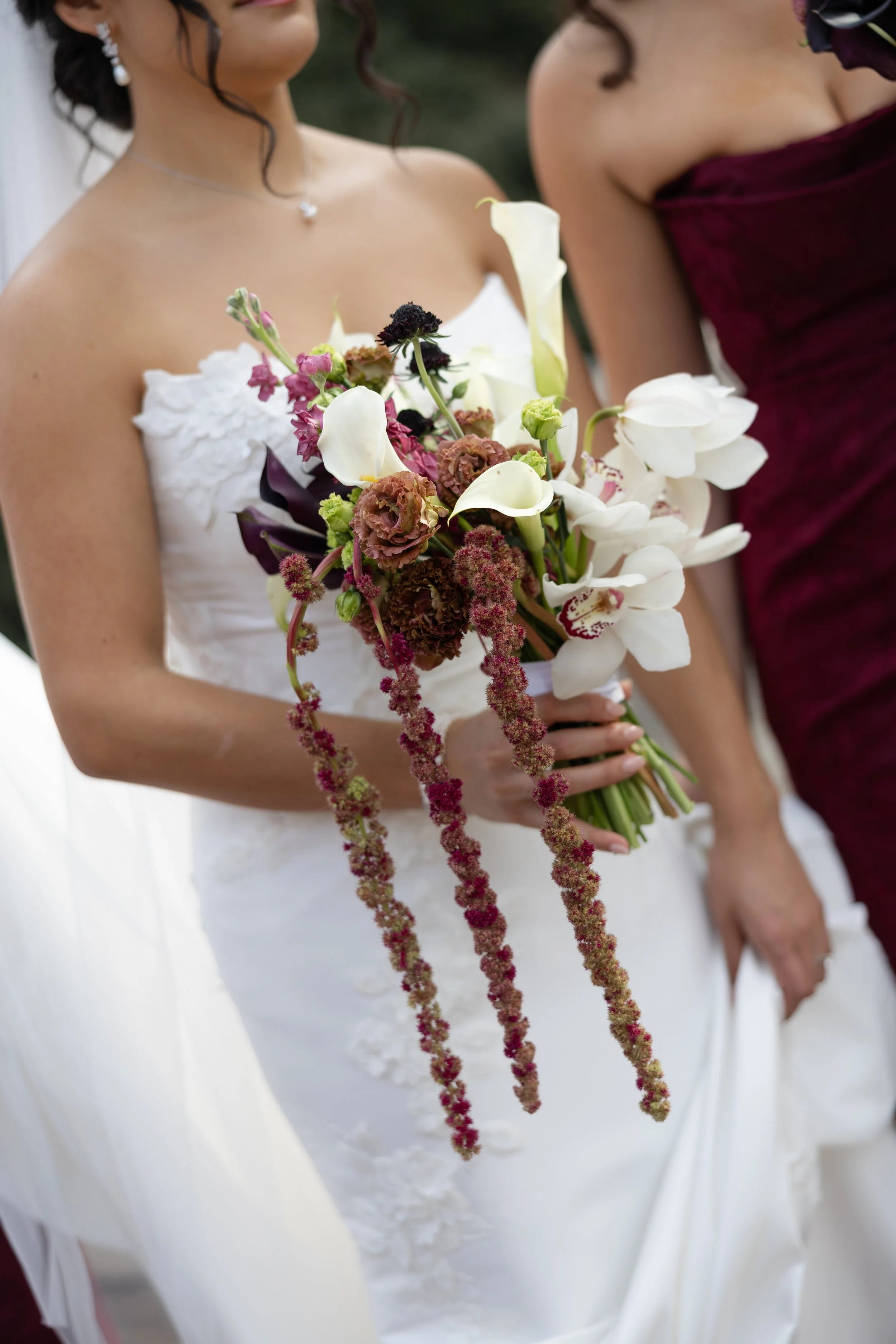 Close-up of a bride holding a bouquet of mixed flowers, with a bridesmaid standing next to her holding her hand. The bride is wearing a white wedding dress with lace details and a necklace with a small heart-shaped pendant.