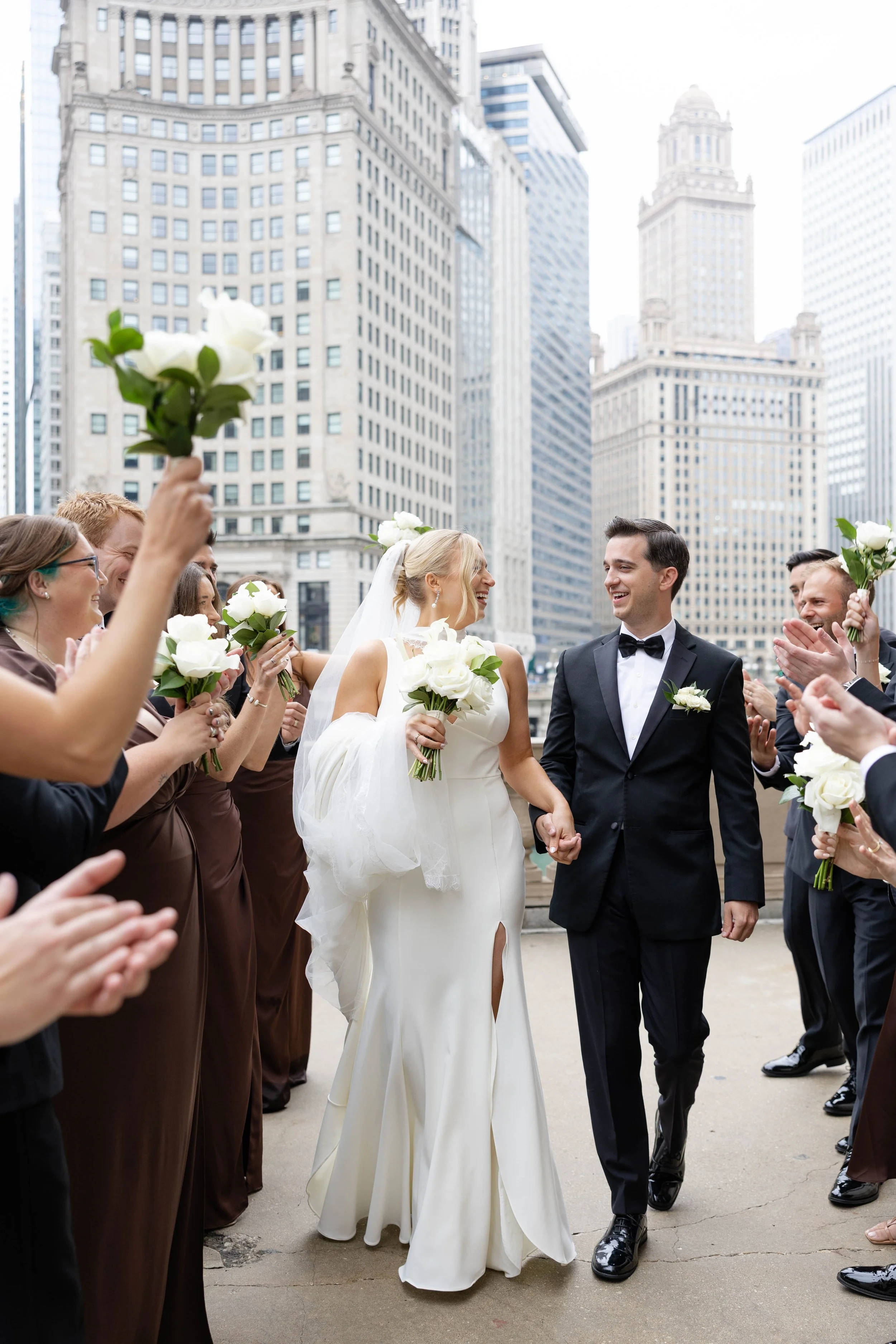 A bride and groom holding hands and smiling at each other during their wedding celebration outdoors in a city, surrounded by friends holding white flowers, with tall buildings in the background.
