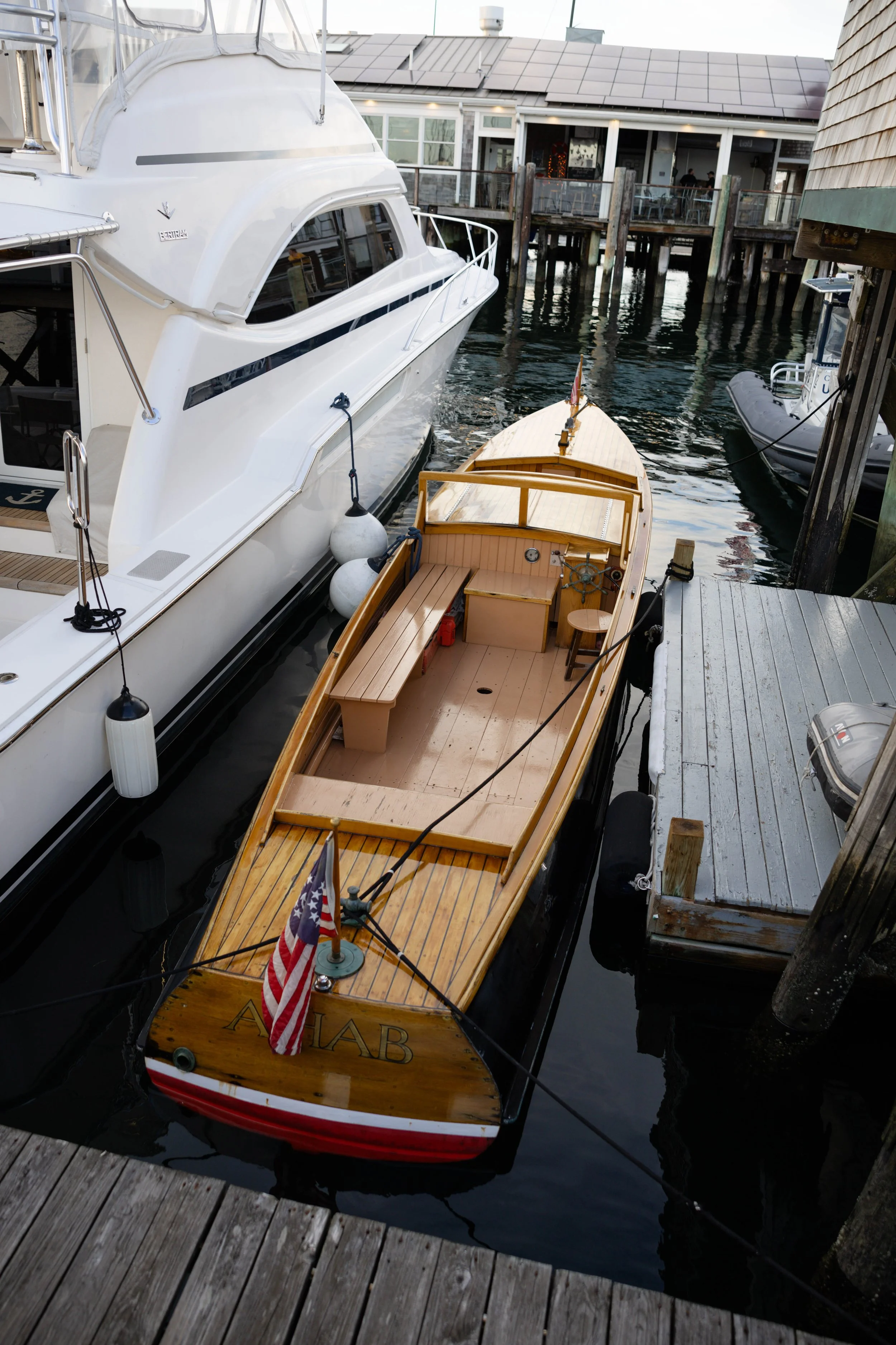 A small wooden boat named 'FLAB' with an American flag docked next to larger white yacht, at a marina with wooden dock and water reflections.