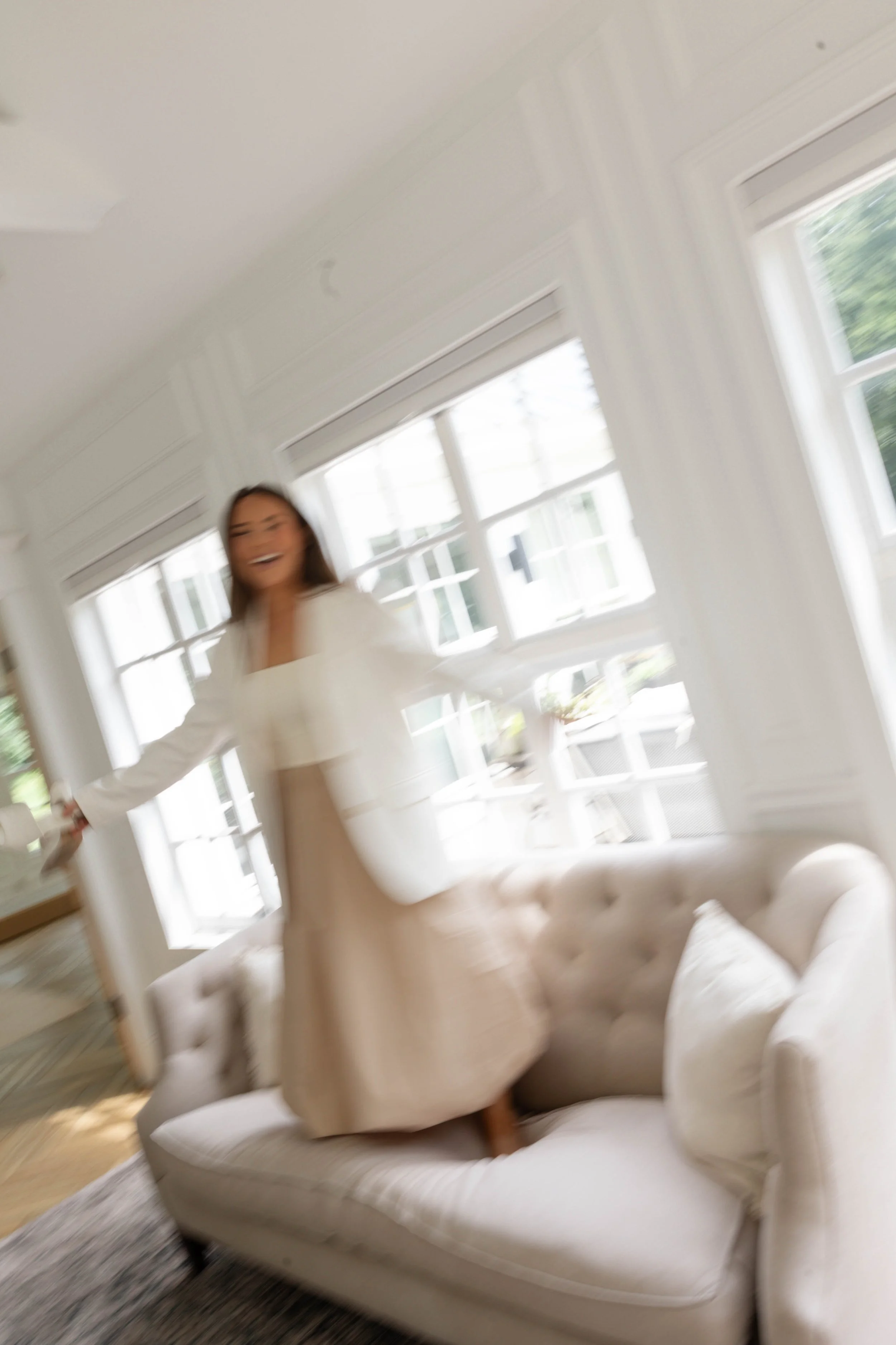 A woman smiling and gesturing inside a bright, white living room with a white tufted sofa, large windows, and a brown wooden floor.