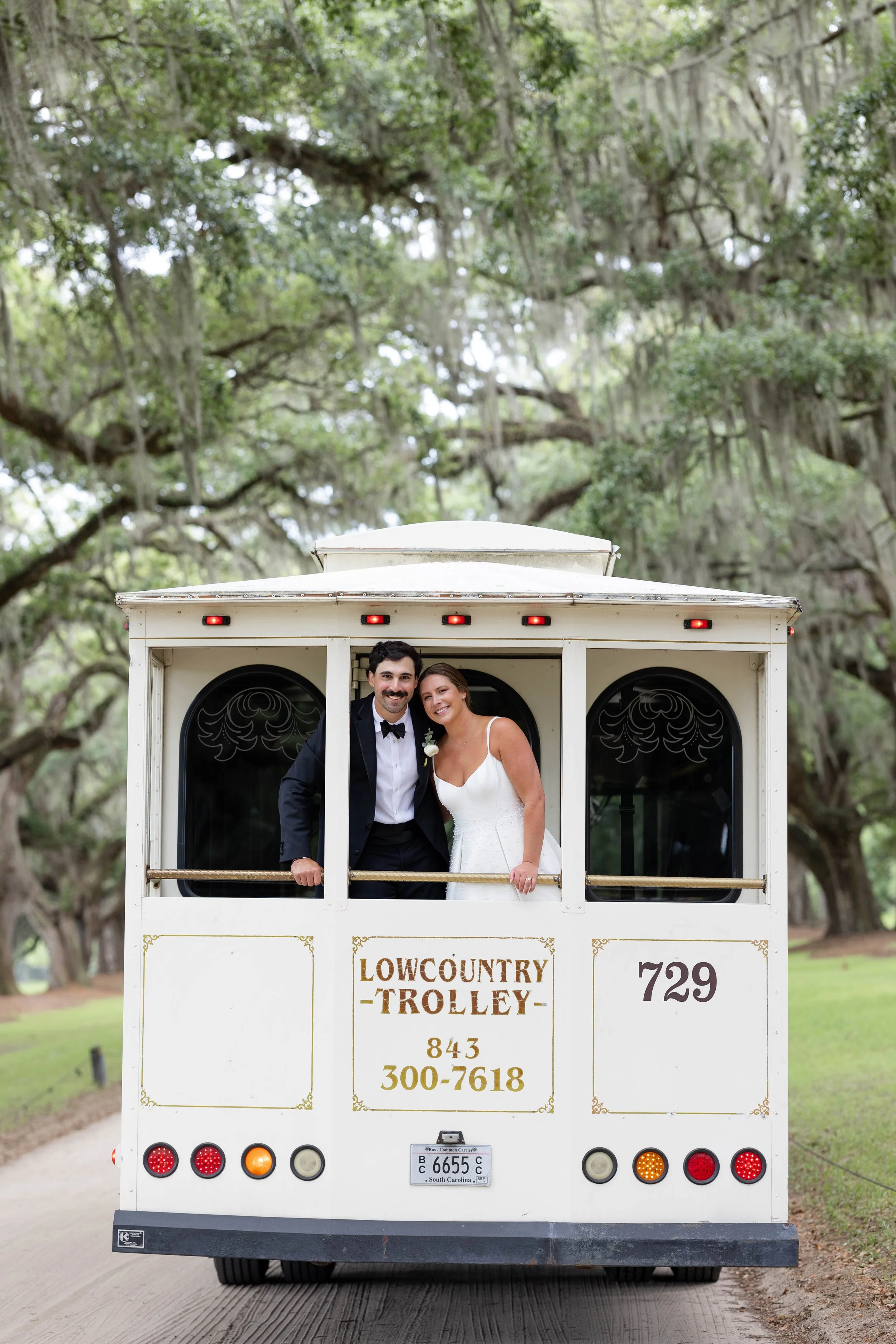 A newlywed couple in wedding attire standing inside a white trolley cart labeled "Lowcountry Trolley" during daytime in a park with large trees and Spanish moss.
