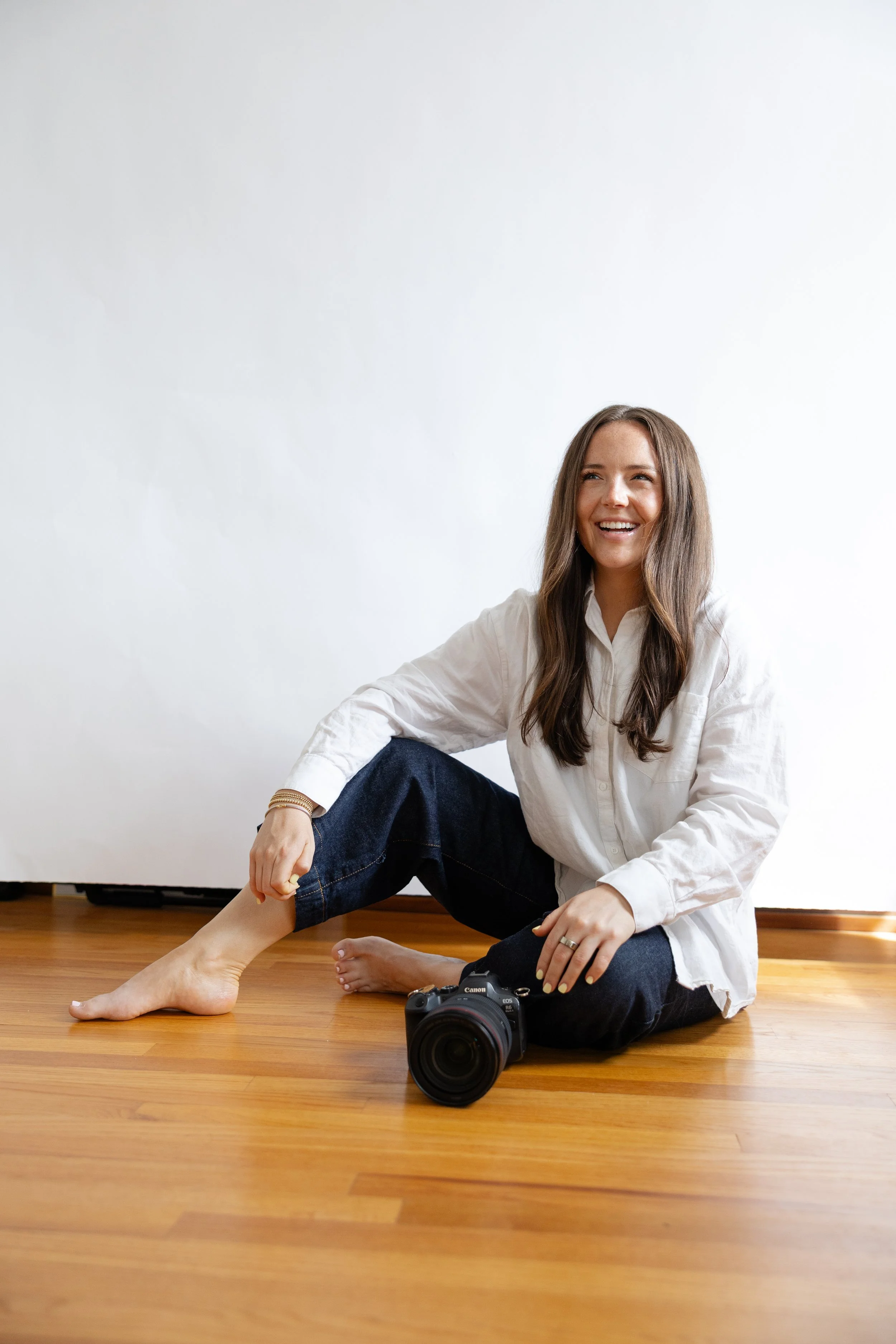 A woman with long brown hair sitting on the wooden floor with a smile, wearing a white shirt and dark jeans, next to a camera, against a plain white wall.
