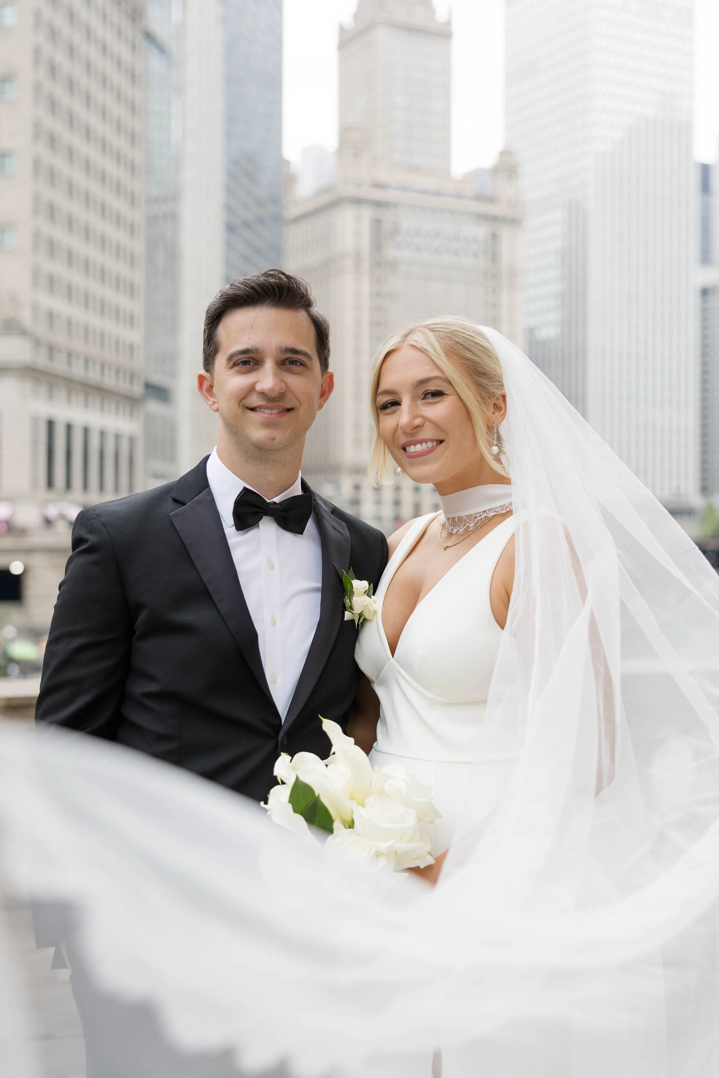 A newlywed couple standing outdoors in front of skyscrapers, with the bride in a white wedding gown and veil and the groom in a black tuxedo, smiling at the camera.