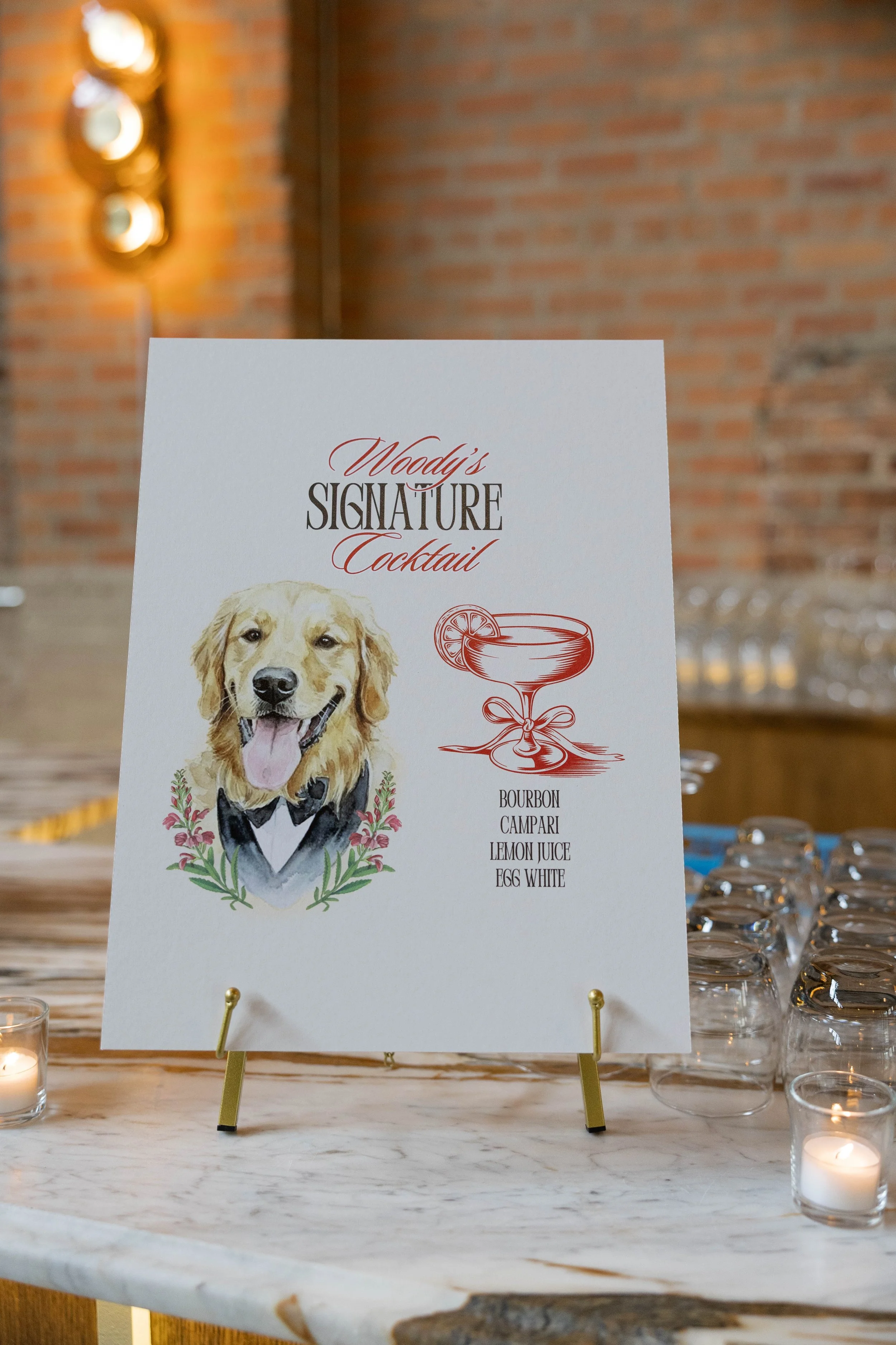 A wedding reception table display featuring a sign with a dog wearing a tuxedo, promoting Woody's signature cocktail with ingredients listed below, amidst scattered glasses and lit candles on a marble table.