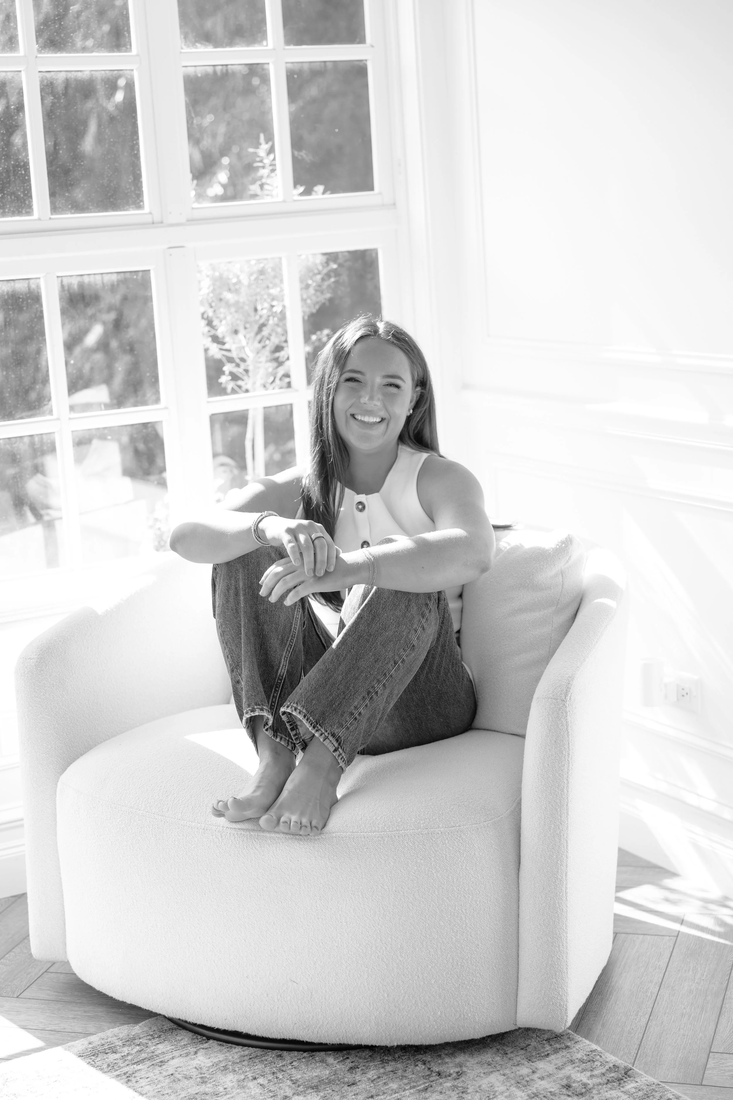 A woman sitting cross-legged on a round, white armchair near a large window, smiling at the camera in a bright room with hardwood floors and a putty-colored rug.