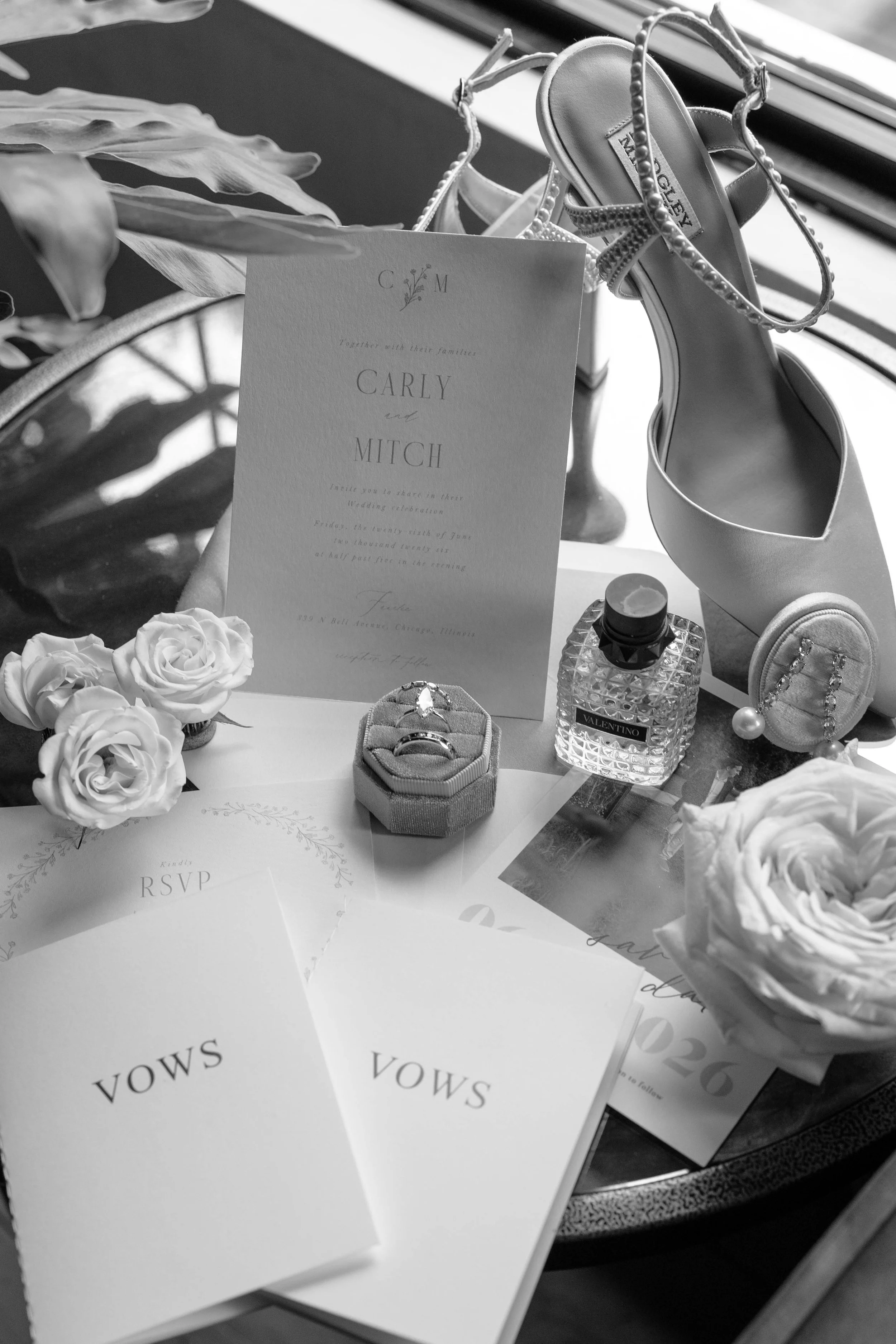 A table with wedding-related items including a white invitation card with the names 'Carly and Mitch,' a small crown-shaped jewelry box, a bottle of Valentino perfume, a pair of white high-heeled shoes with pearl embellishments, white roses, and RSVP