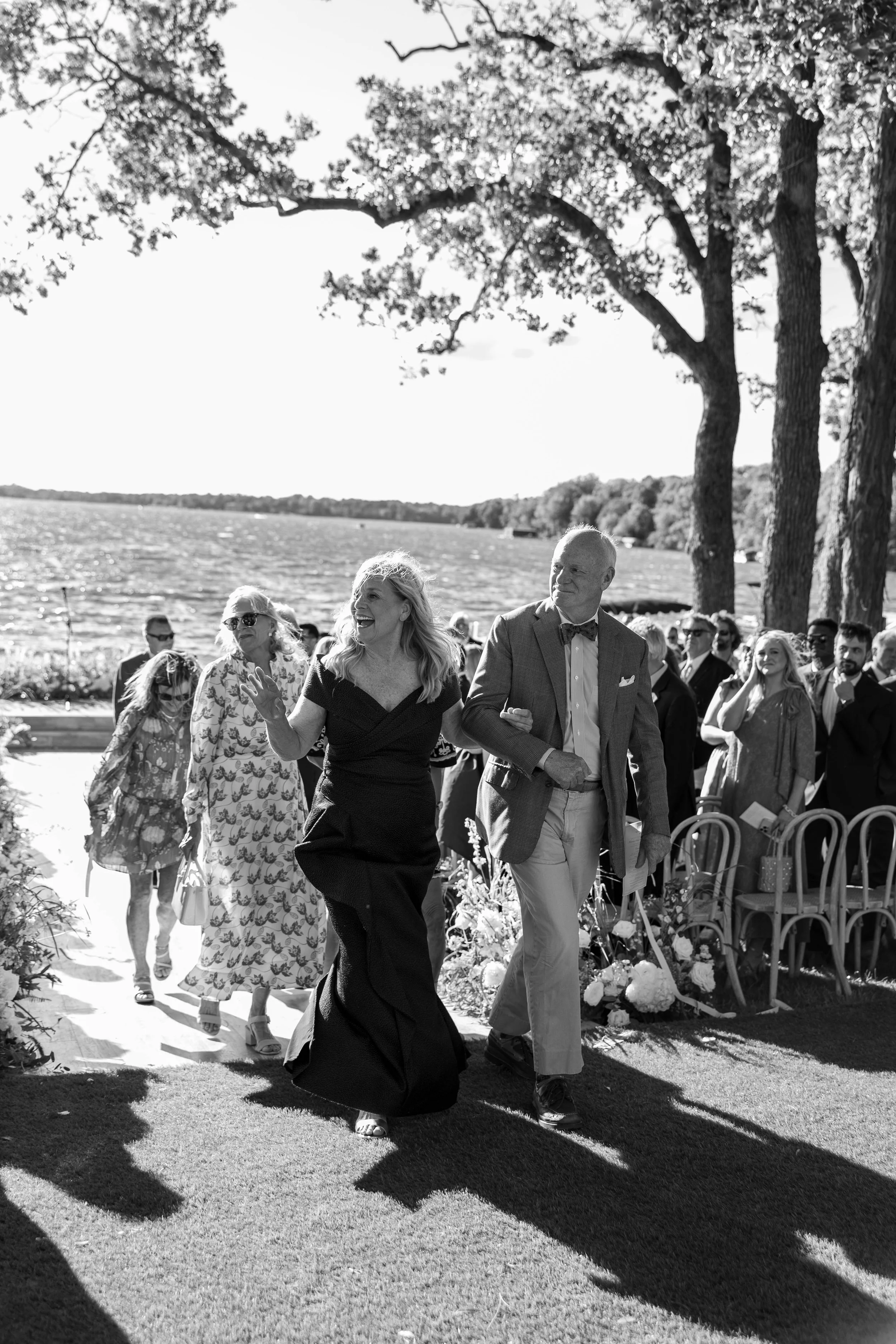 People dressed in formal attire walking outdoors near a lake with trees in the background during daytime.