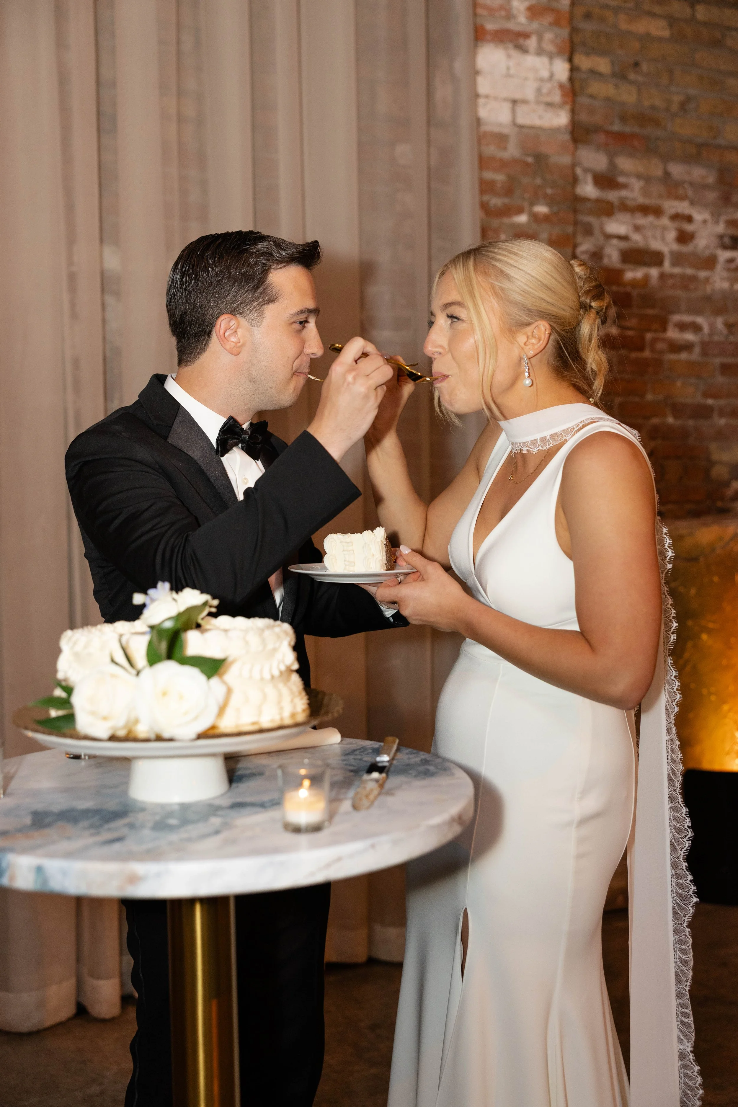 A bride and groom at their wedding reception sharing a spoonful of cake. The groom is in a black tuxedo, and the bride is in a white wedding dress. There is a wedding cake with flowers on a table in the foreground.