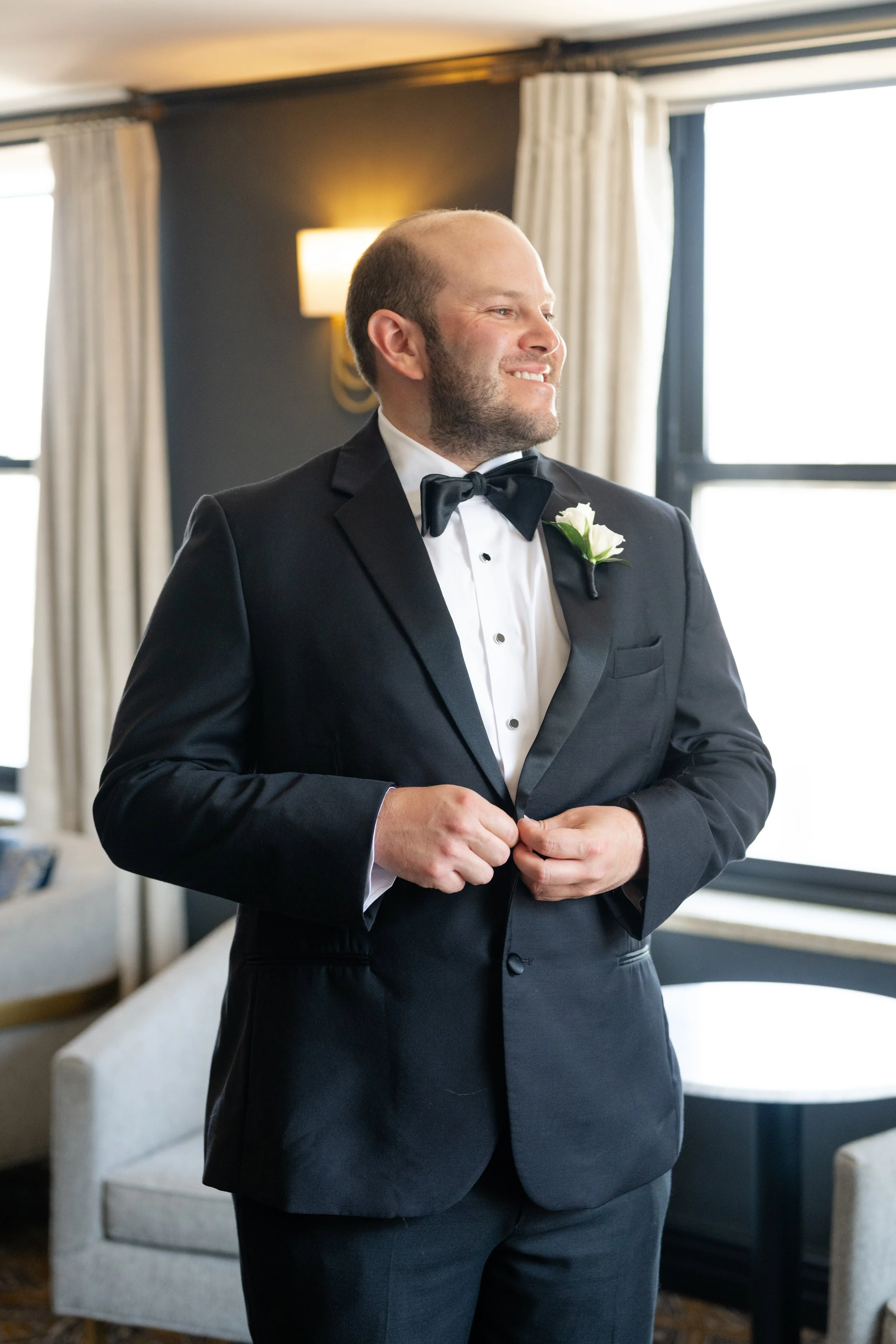 A man in a tuxedo with a white boutonniere smiles and adjusts his jacket in a warmly lit room with large windows, cream curtains, and modern decor.