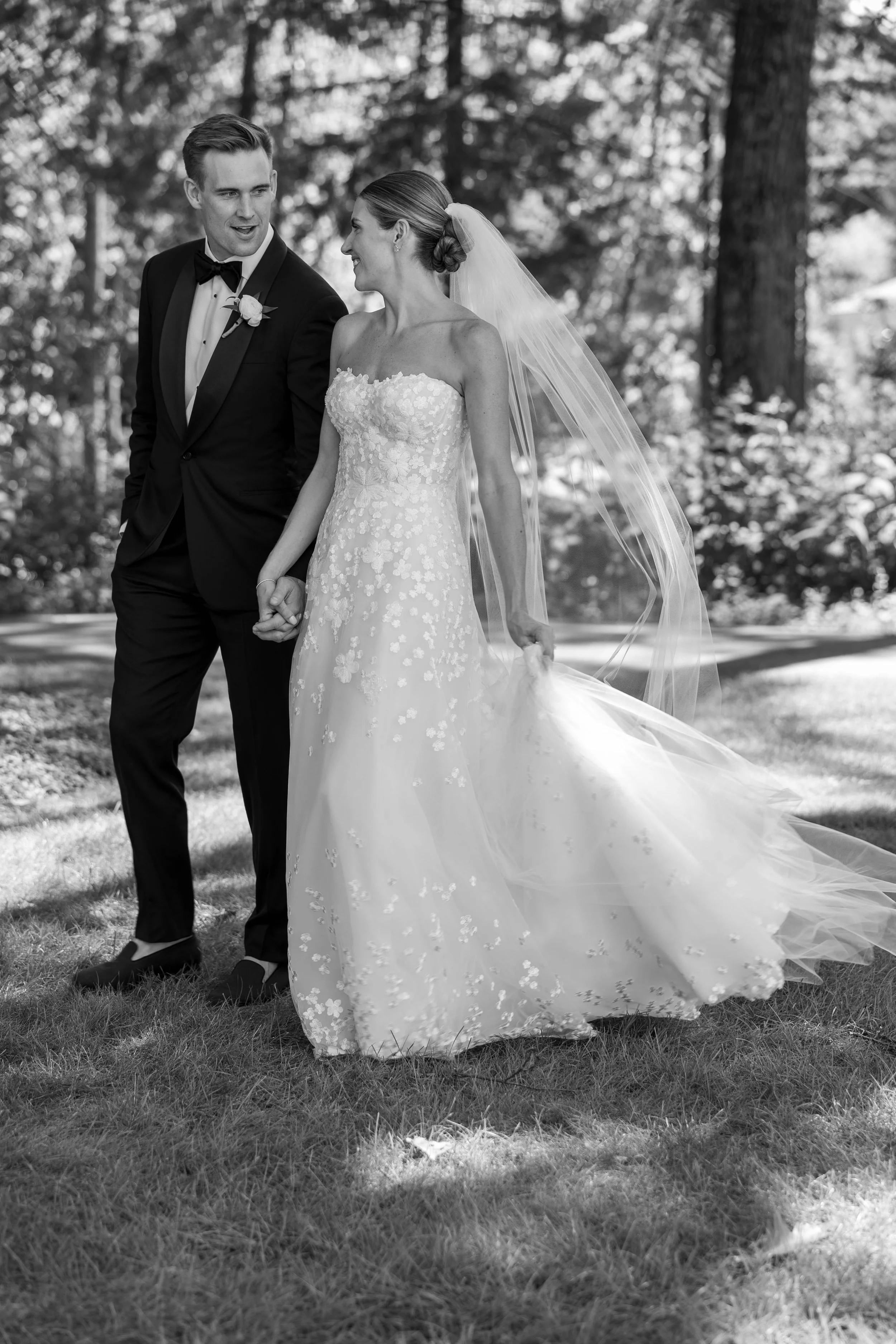 A black-and-white photo of a bride and groom holding hands outdoors, walking on grass in a wooded area, dressed in wedding attire. The groom wears a tuxedo with a bow tie, and the bride wears a strapless wedding gown with floral details and a long veil.