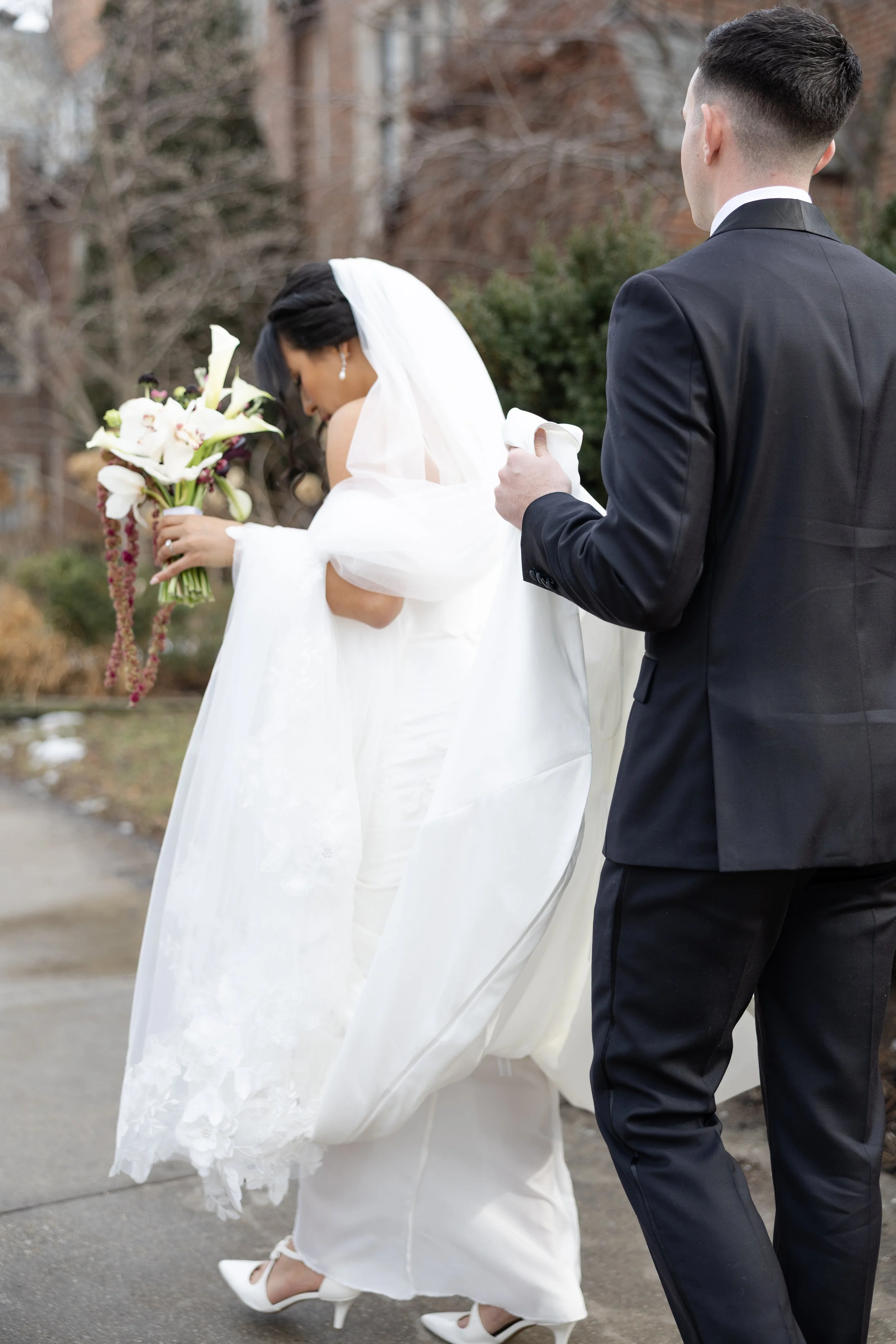 A bride in a white wedding gown holding a bouquet of white lilies and orchids, walking with a groom in a black suit, outdoors during daytime. The bride is looking down, and the groom is holding her train, facing away from the camera.