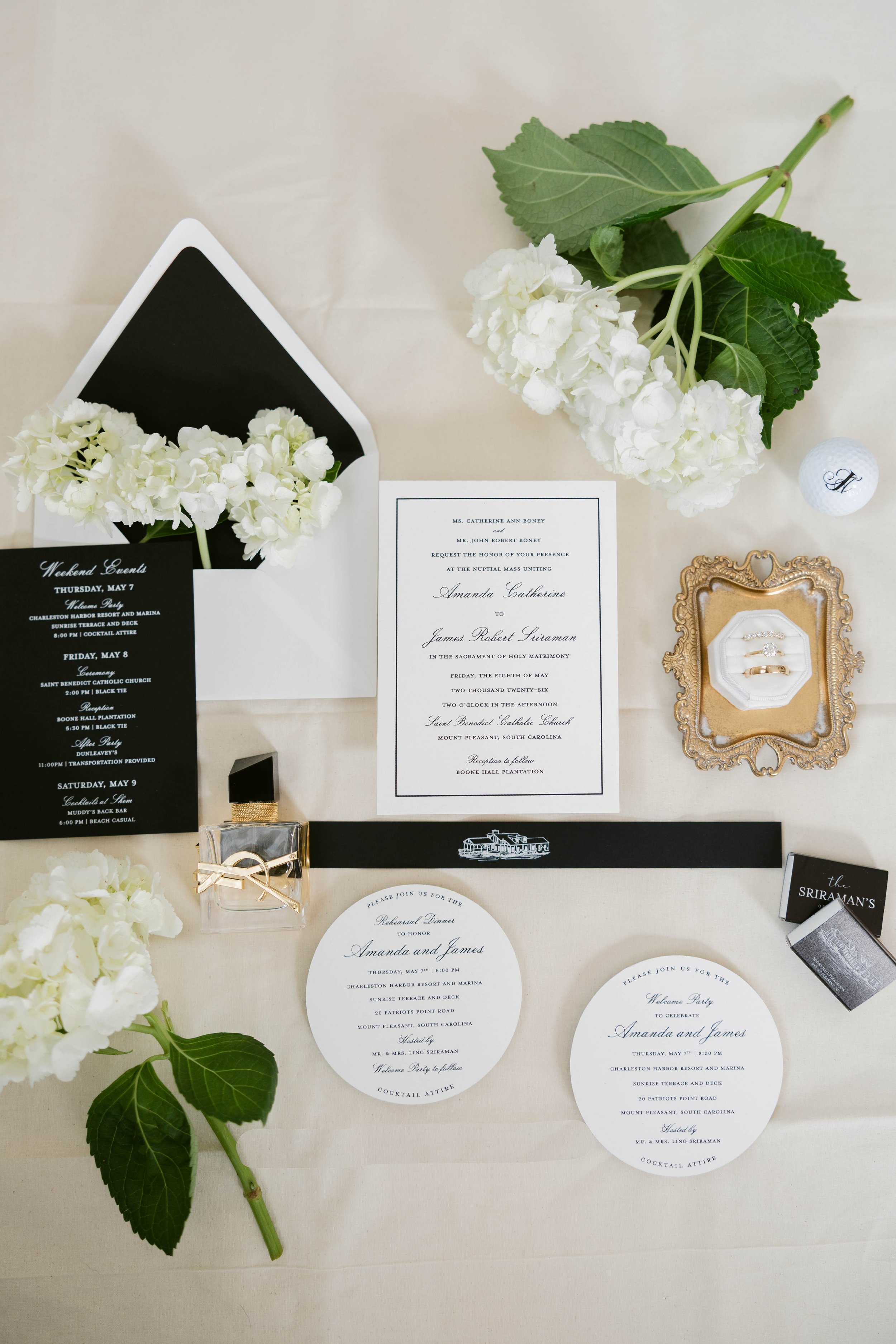 Wedding invitation suite, white hydrangea flowers, a golf ball with a letter 'A', and gold wedding rings in a decorative box on a cream tablecloth.